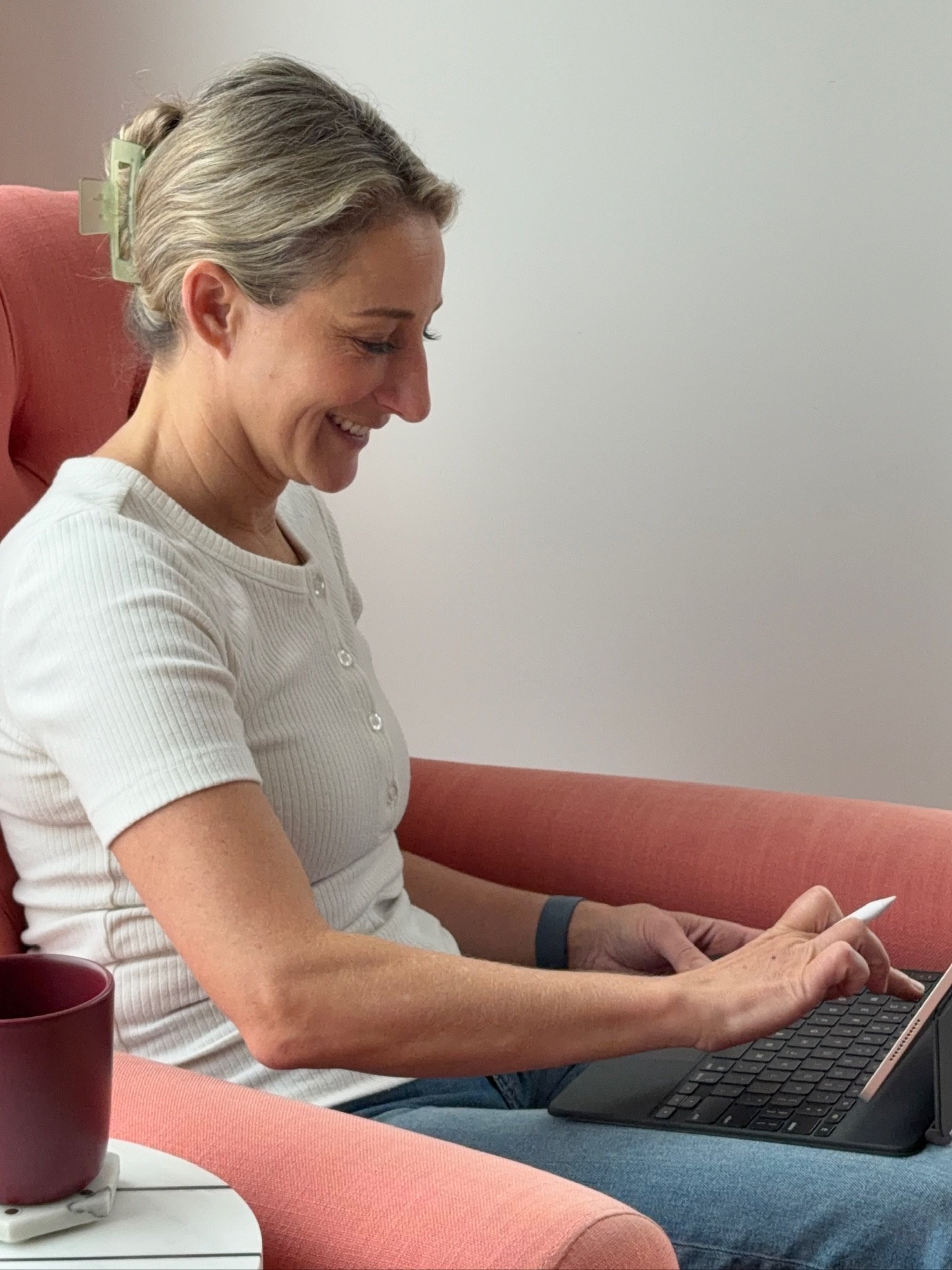 A woman with gray hair tied with a green hair clip, wearing a white shirt, sitting on a red chair, smiling, and typing on a laptop. There is a red mug on a small table next to her.