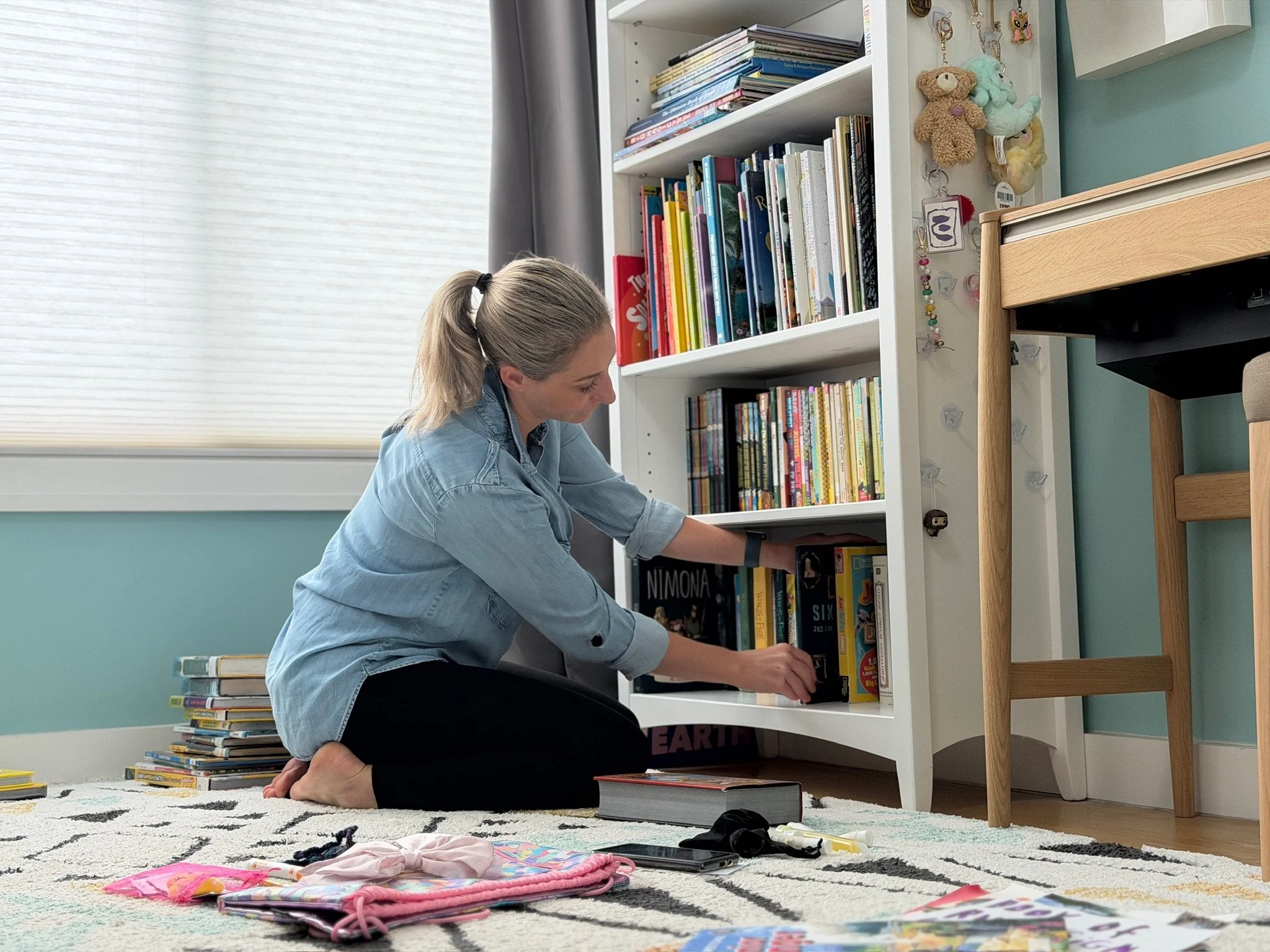 A woman with blonde hair tied back, sitting on the floor and reading a book. She is wearing a denim shirt and black pants. Behind her, there is a white bookshelf filled with various books and some stuffed animals on top. The room has a light blue wall, a window with closed blinds, and a patterned rug on the floor.