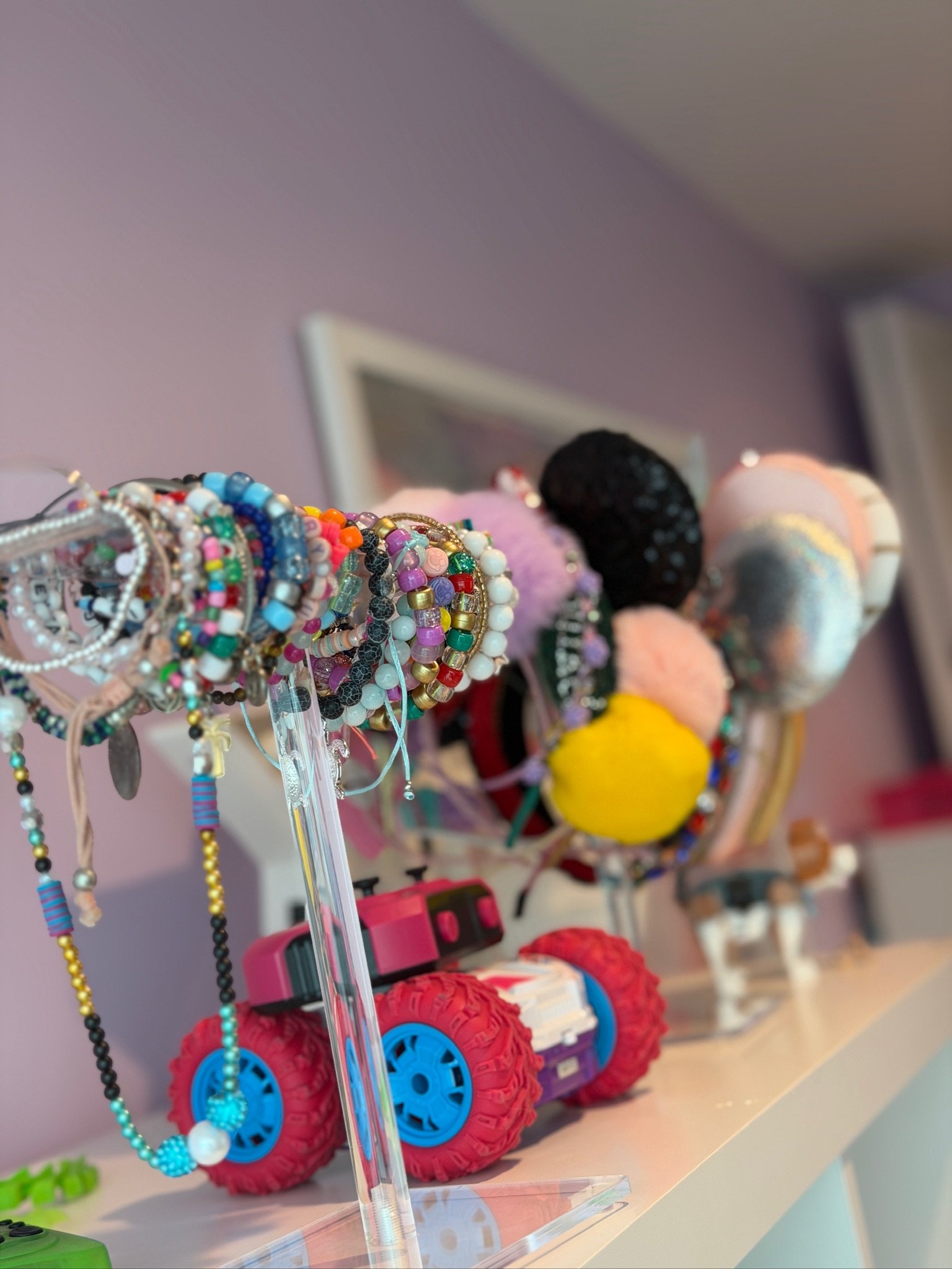 Display of colorful beaded bracelets and jewelry on a stand, with pink and blue toy wheels in the background.