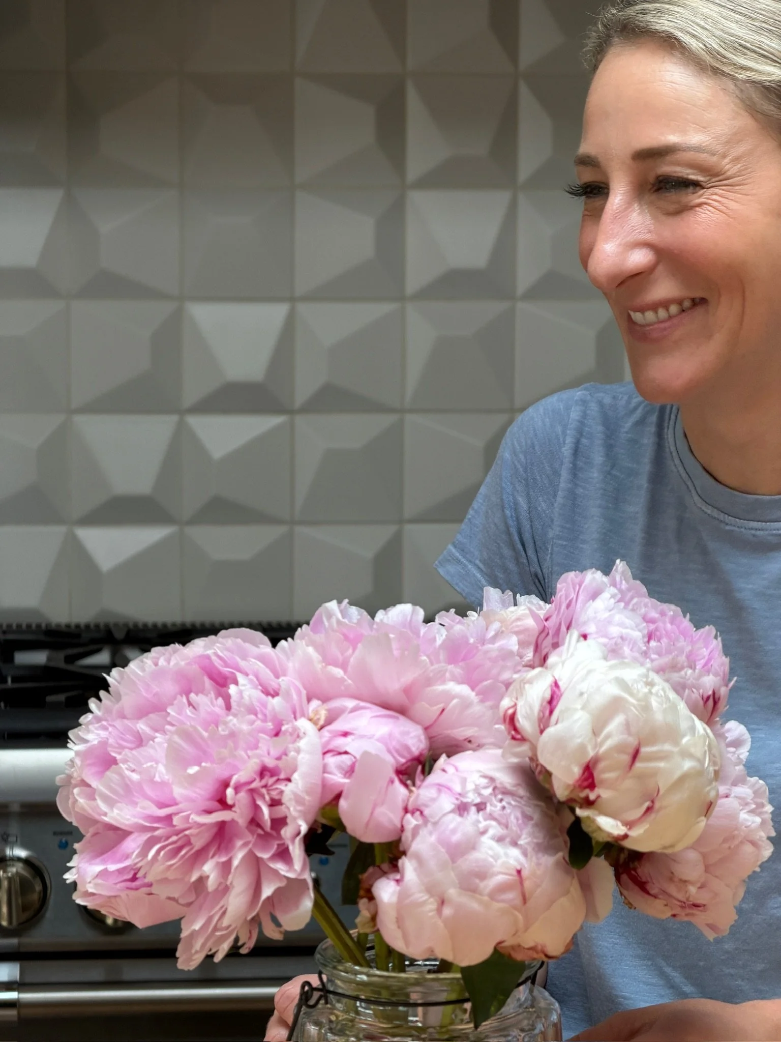 A woman holding a jar of pink and white peonies in front of a tiled wall and stove.