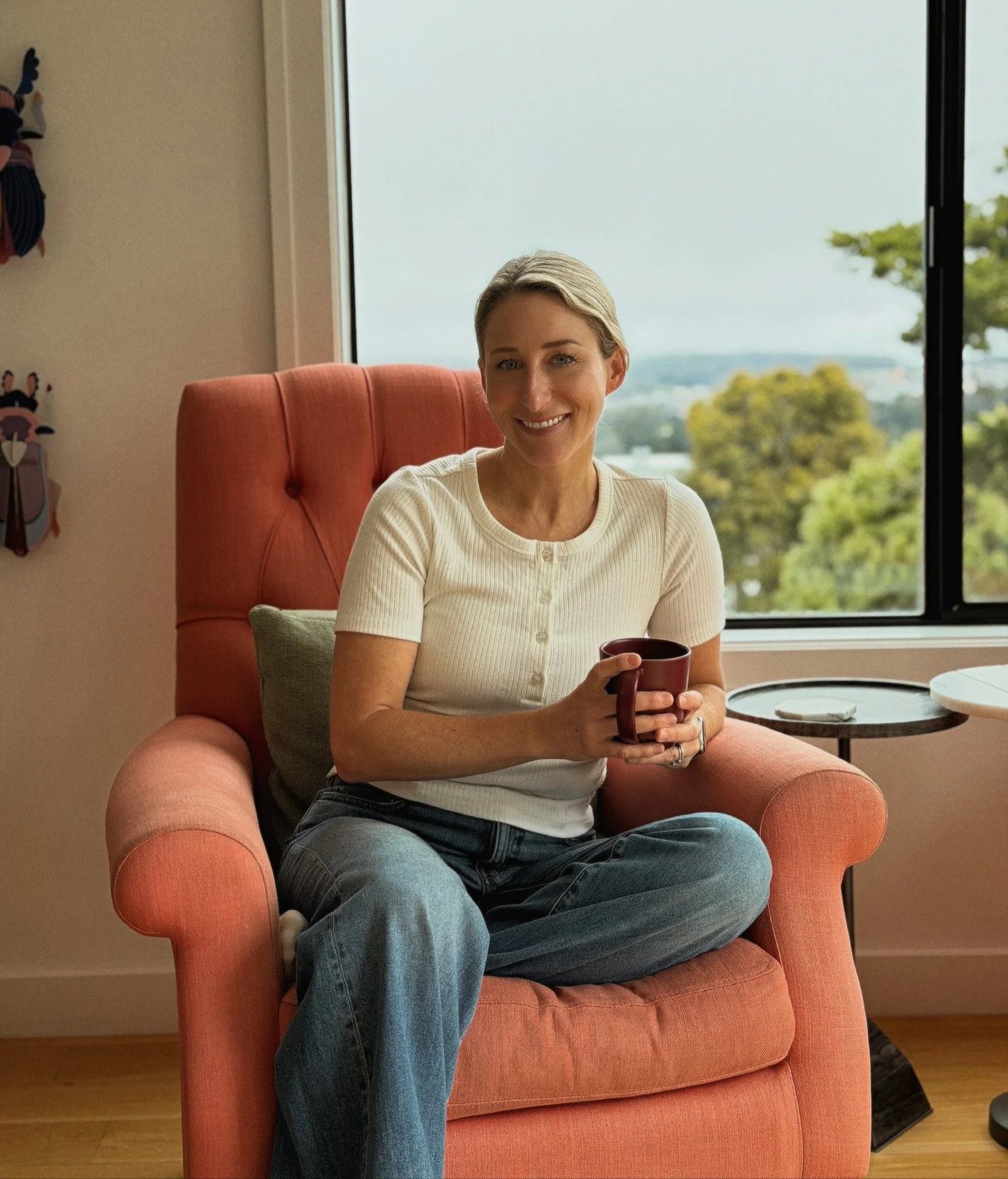 A young woman with blonde hair, sitting on an orange armchair, smiling and holding a mug in both hands. She is wearing a cream-colored, short-sleeved, button-up shirt and jeans. Behind her, there is a large window showing greenery and a partly cloudy sky. A small side table with a smartphone and a dish is next to her.