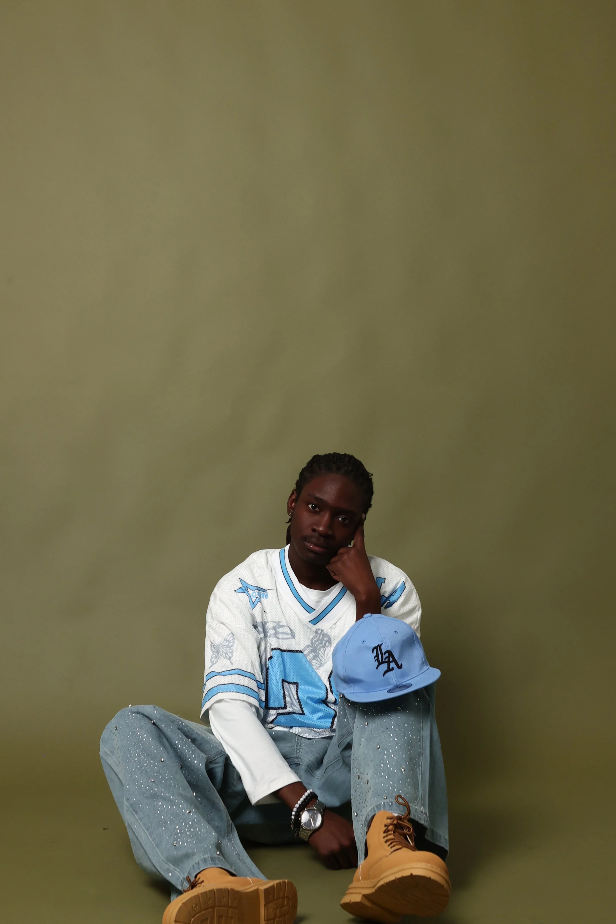 A young man with dark skin, braids, and wearing a white and blue sports jersey, gray distressed jeans, and tan work boots, sitting on the floor against a plain green wall. He is resting his head on his hand, looking at the camera.