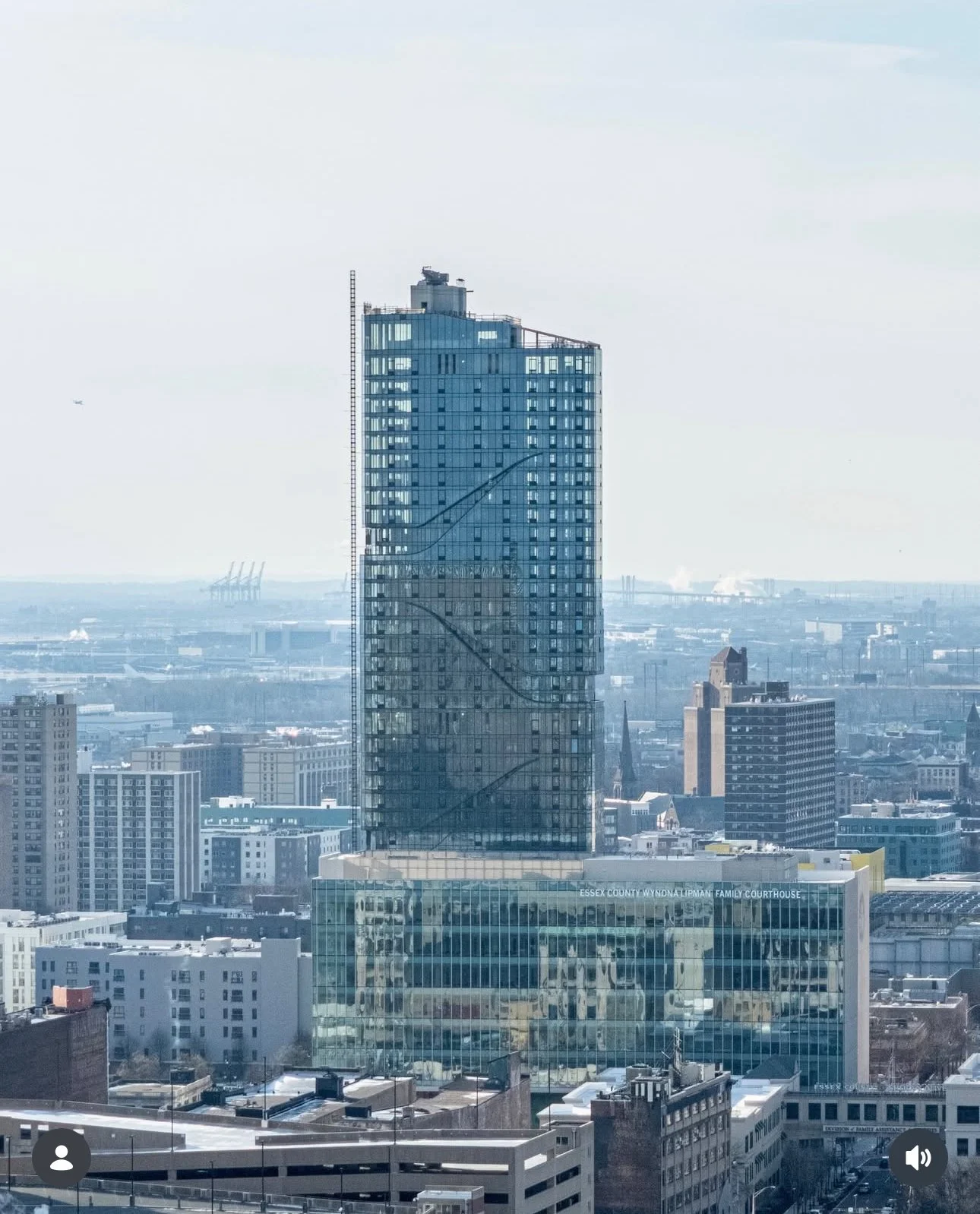 A tall modern glass skyscraper in a cityscape with other buildings, industrial structures, and a clear sky in the background.