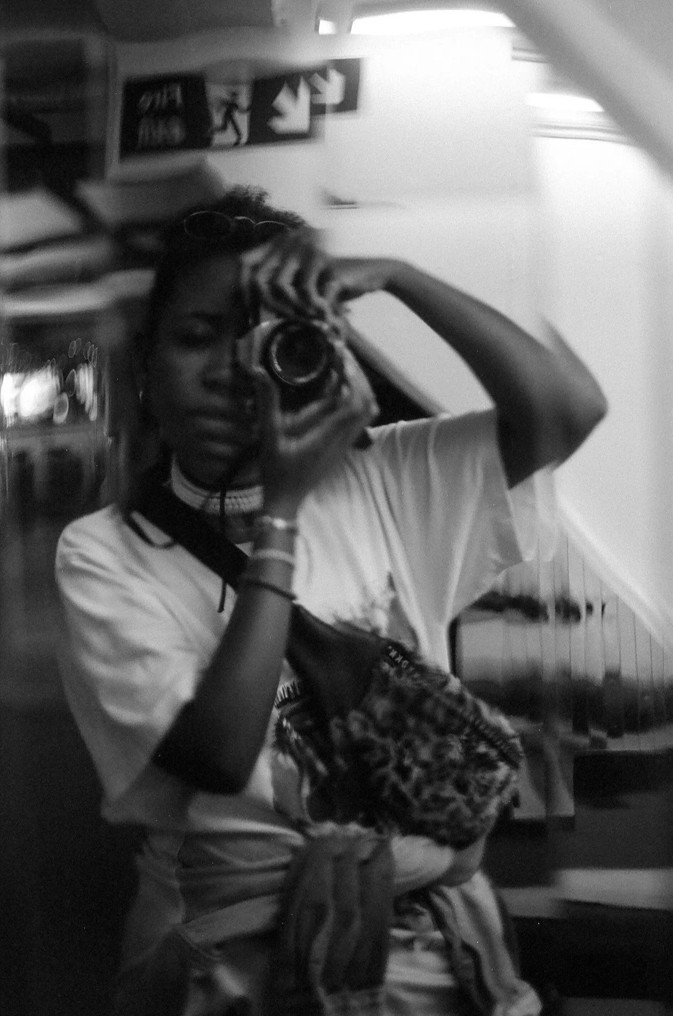 A black-and-white self-portrait of Elena Bueriberi while in London, looking into the mirror inside of a building while holding her Canon AE-1 film camera.