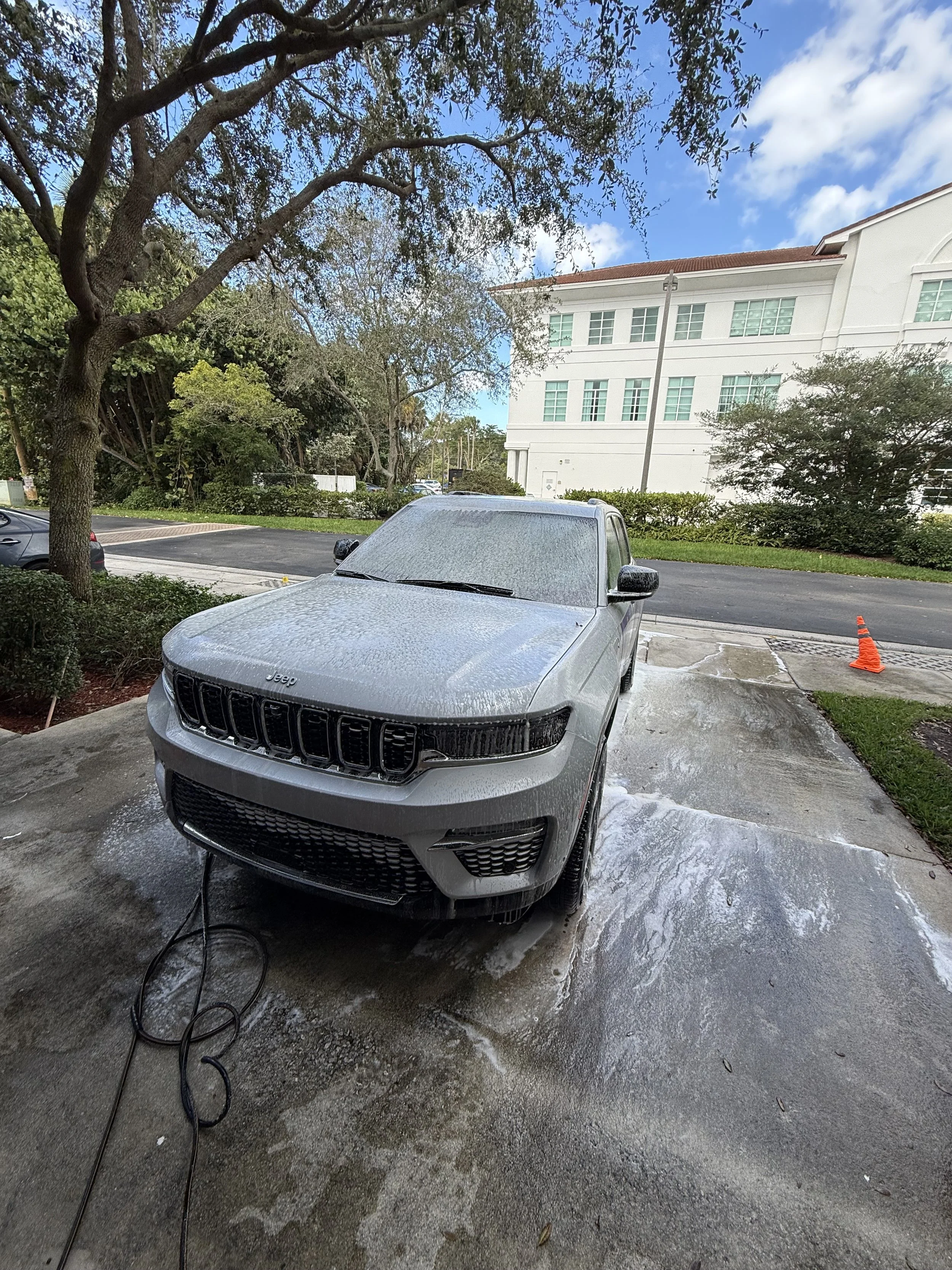 A gray Jeep SUV is covered in soap suds during a car wash, with a wet concrete surface and an orange cone nearby, in an outdoor parking lot with trees and a white building in the background.