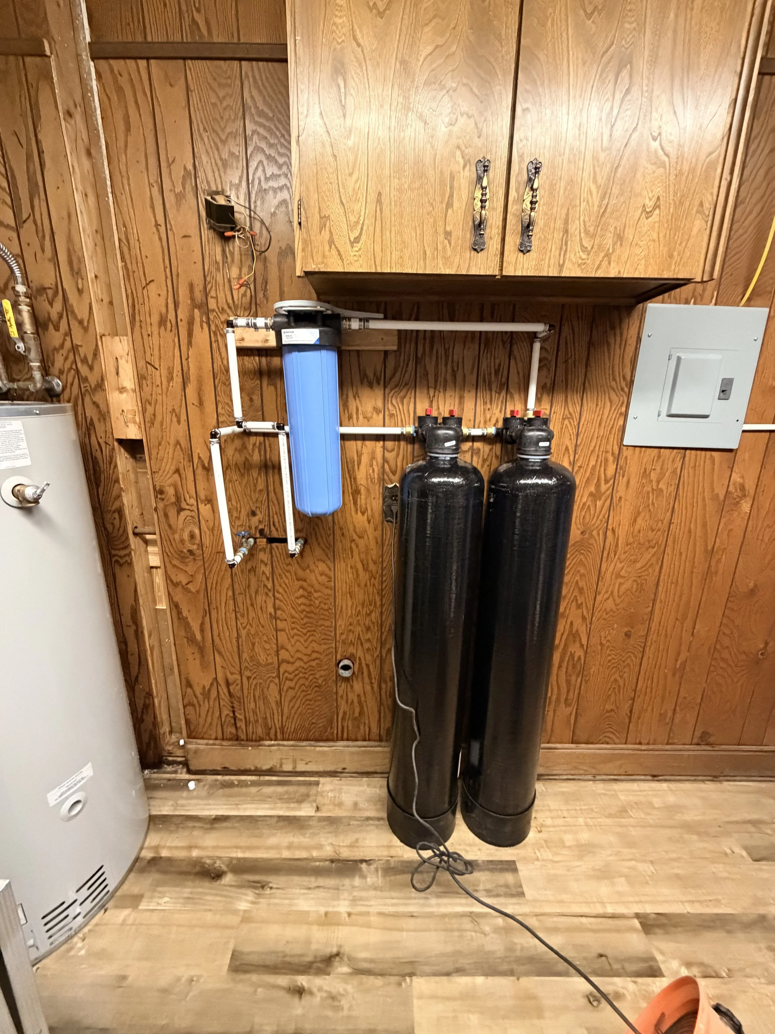 Hoses, water filter system, and water softener tanks in a utility room with wooden paneling.