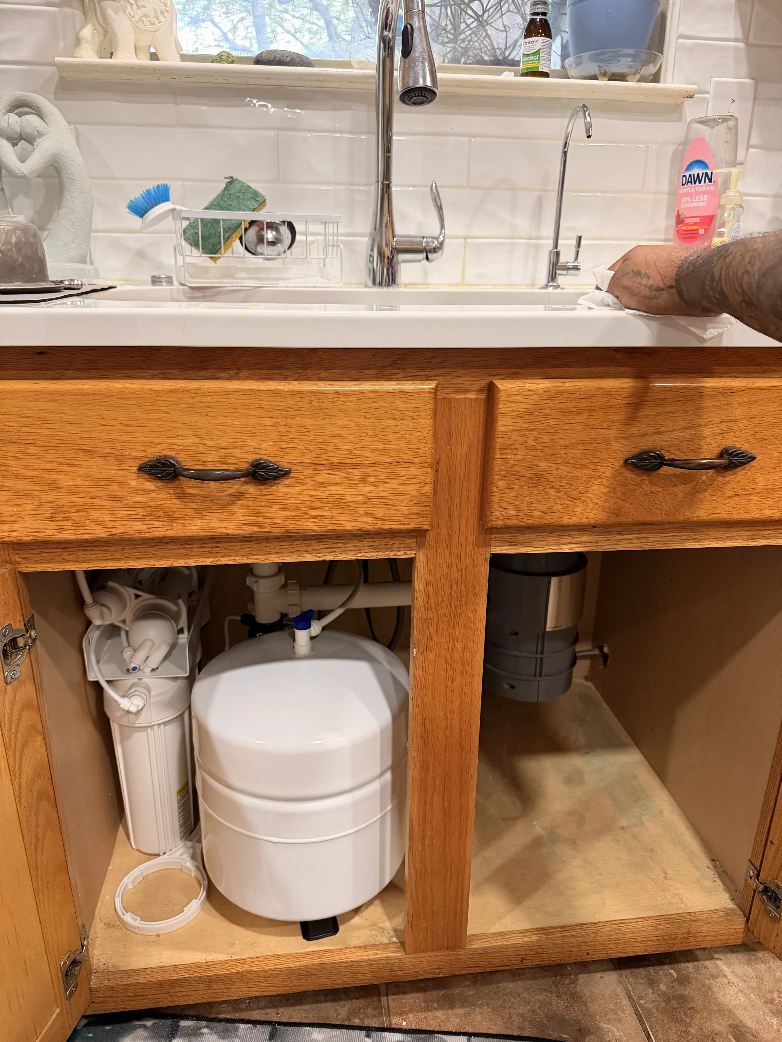 Inside a kitchen cabinet under the sink, with plumbing and a water filter. The cabinet door is open, revealing the water filter system and some plumbing pipes. Someone is cleaning the countertop with a paper towel.