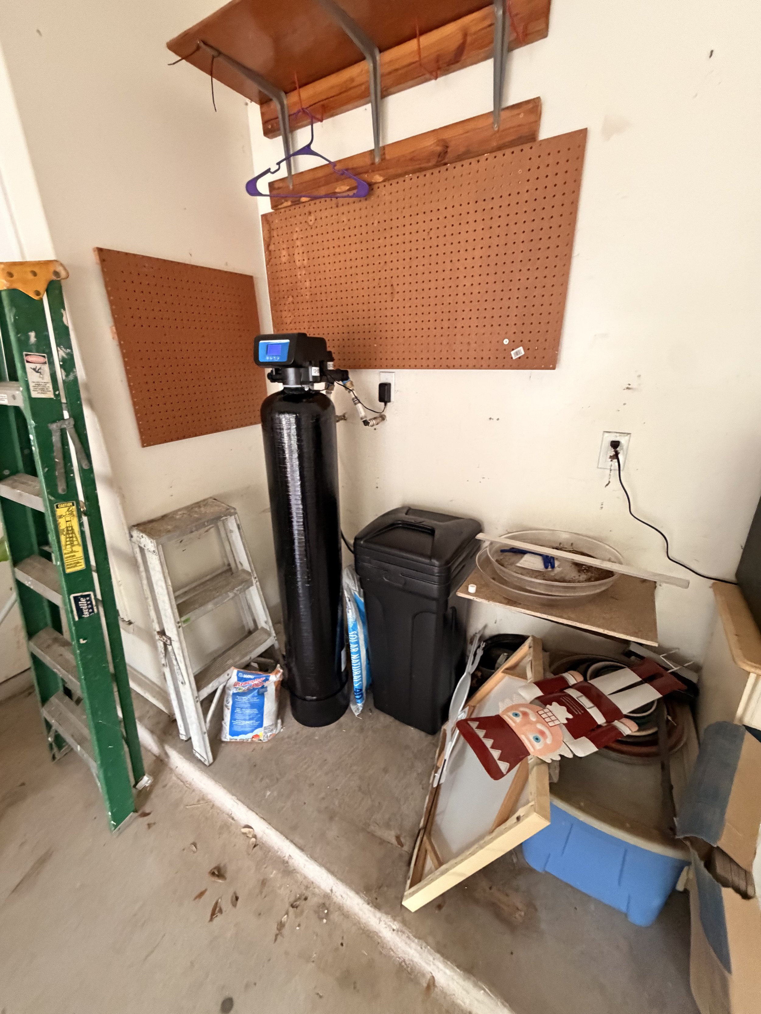 A cluttered garage corner with a ladder, two black water softener tanks, a trash bin, and a pegboard on the wall, with various tools and items on and around a workbench.