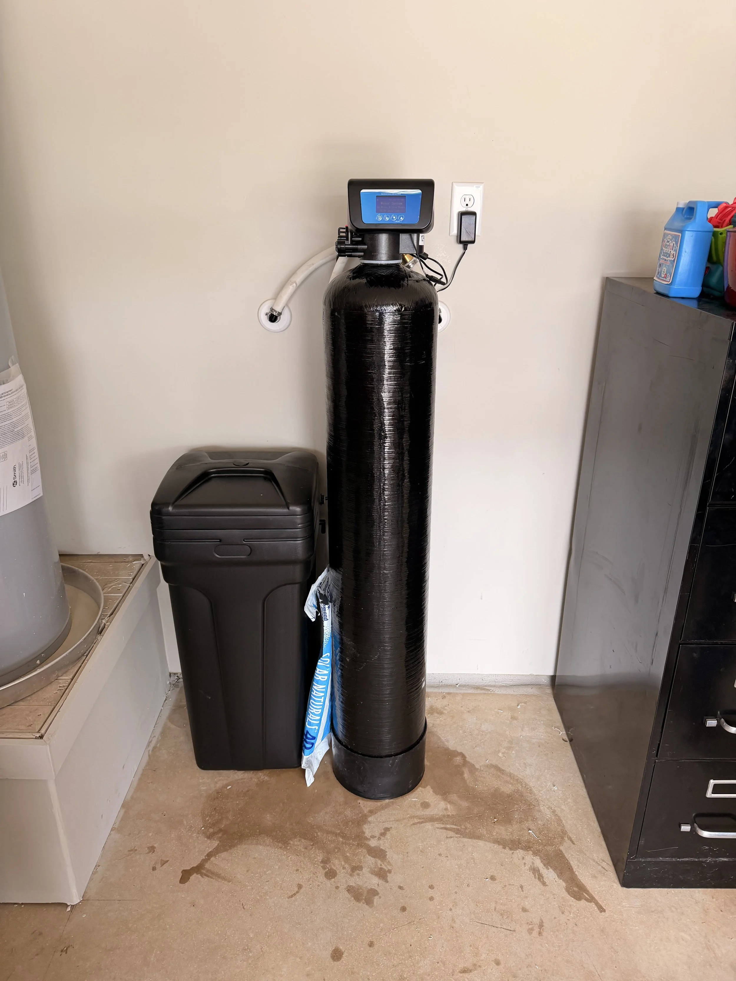 Water softener with digital control panel, connected to water piping, placed next to a black trash can in utility room.