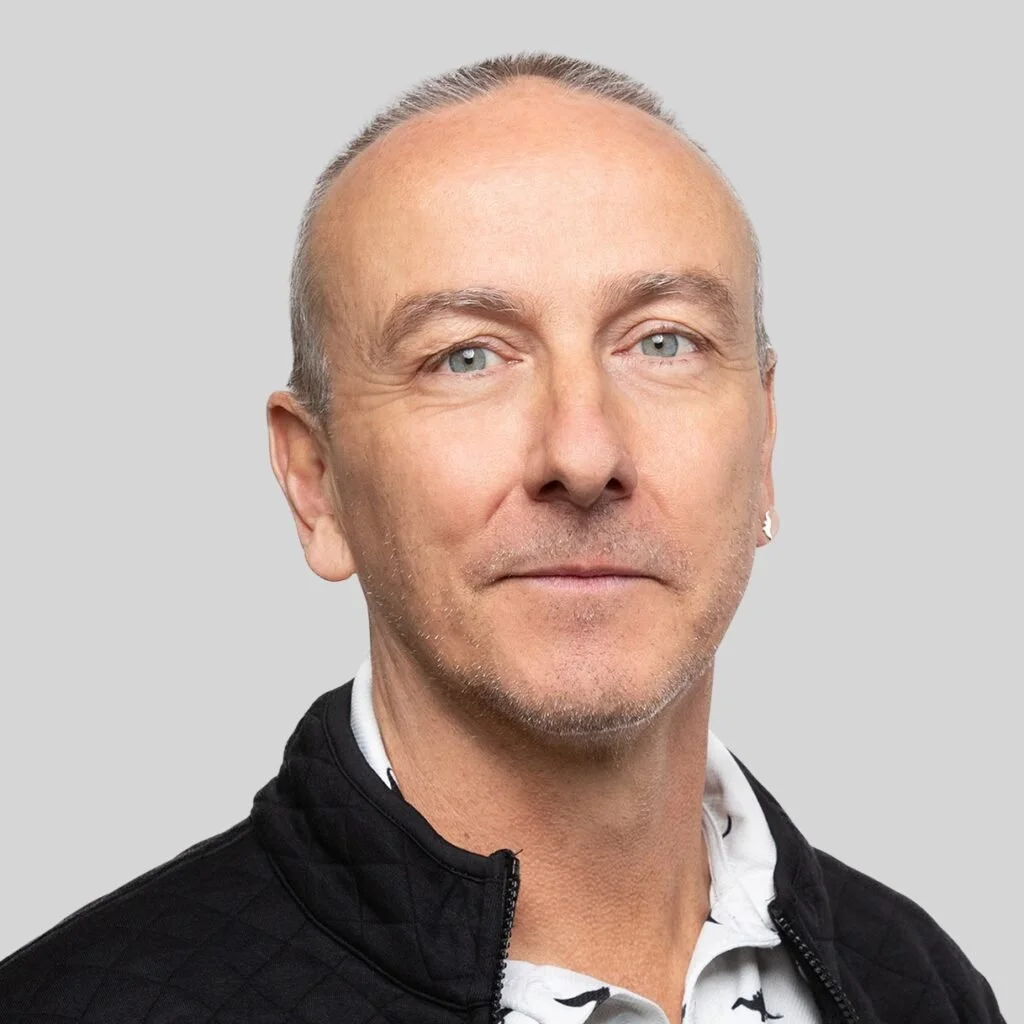 Close-up portrait of a middle-aged man with short, light-colored hair, blue eyes, and a slight smile, wearing a black jacket over a white shirt with a pattern.