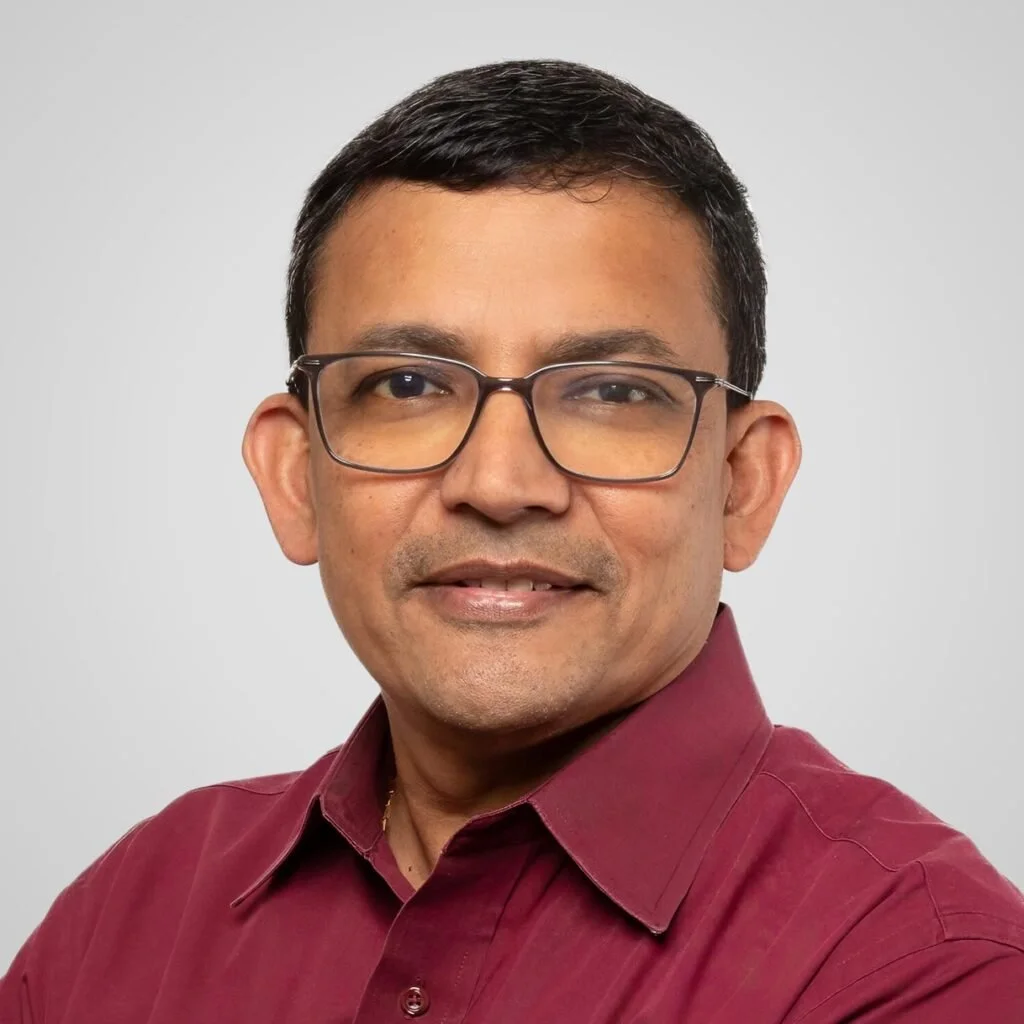 Professional headshot of a man with short black hair, wearing glasses and a maroon collared shirt, smiling slightly against a plain light gray background.