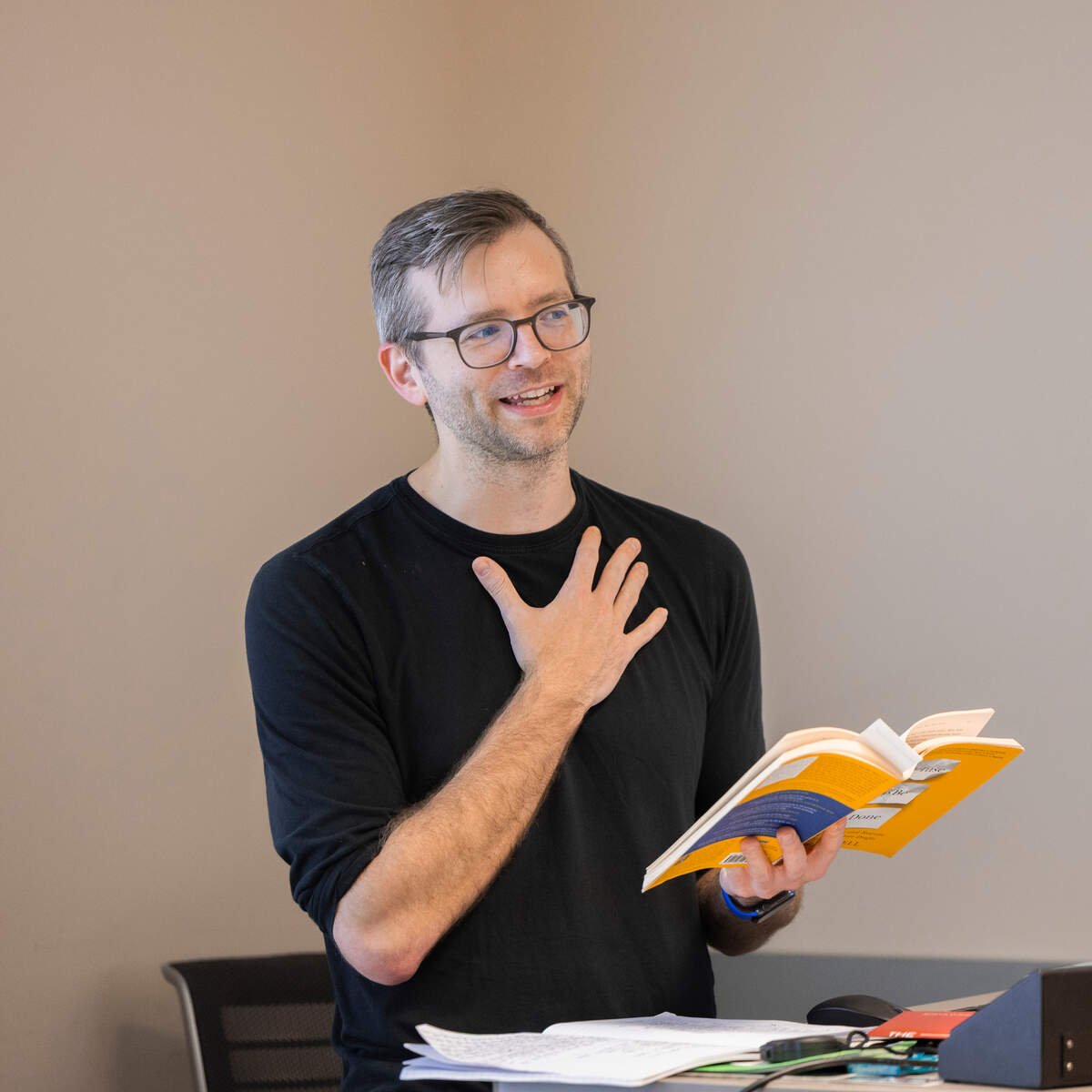 Caleb Tankersly with glasses, wearing a black shirt, is speaking with a hand on his chest while holding an open book in his other hand in a room with a neutral background.