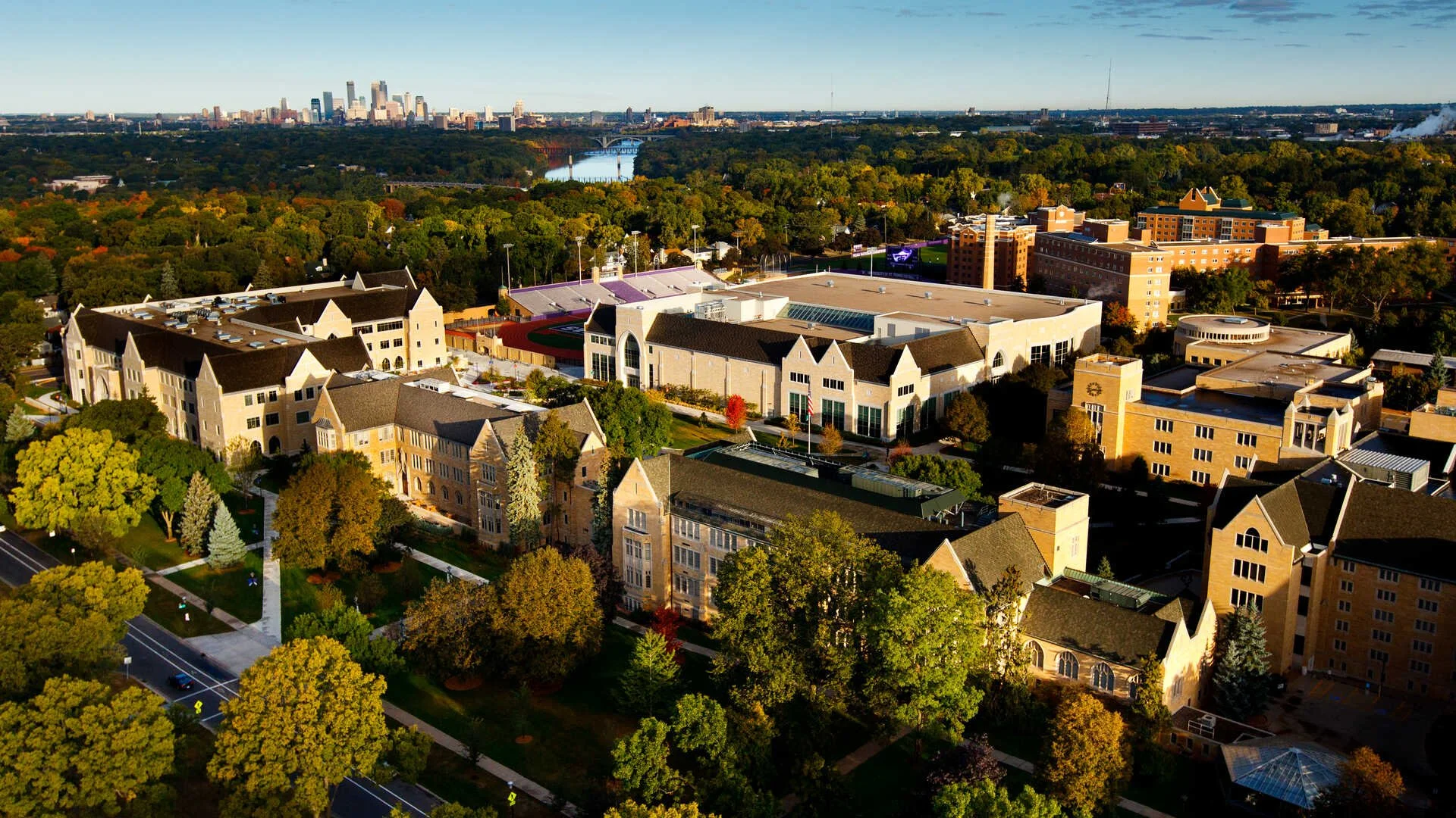Aerial view of University of St. Thomas campus with historic stone buildings, trees, sports fields, and a city skyline in the background.