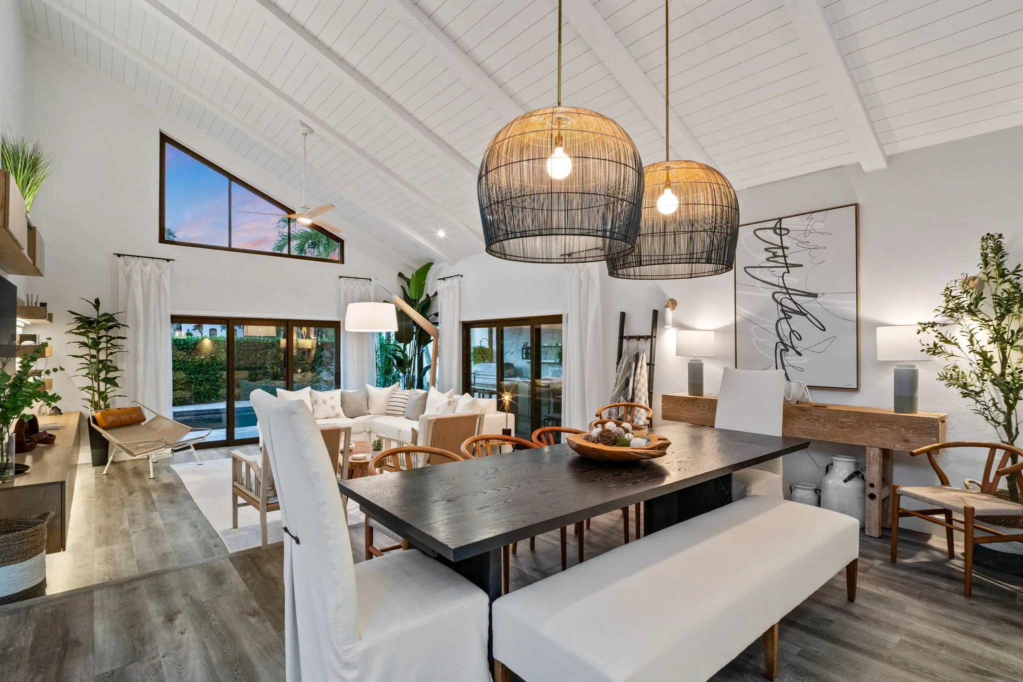 Open concept living and dining room with high white ceiling, large windows, pendant lights, various plants, comfortable seating, and wooden furniture.