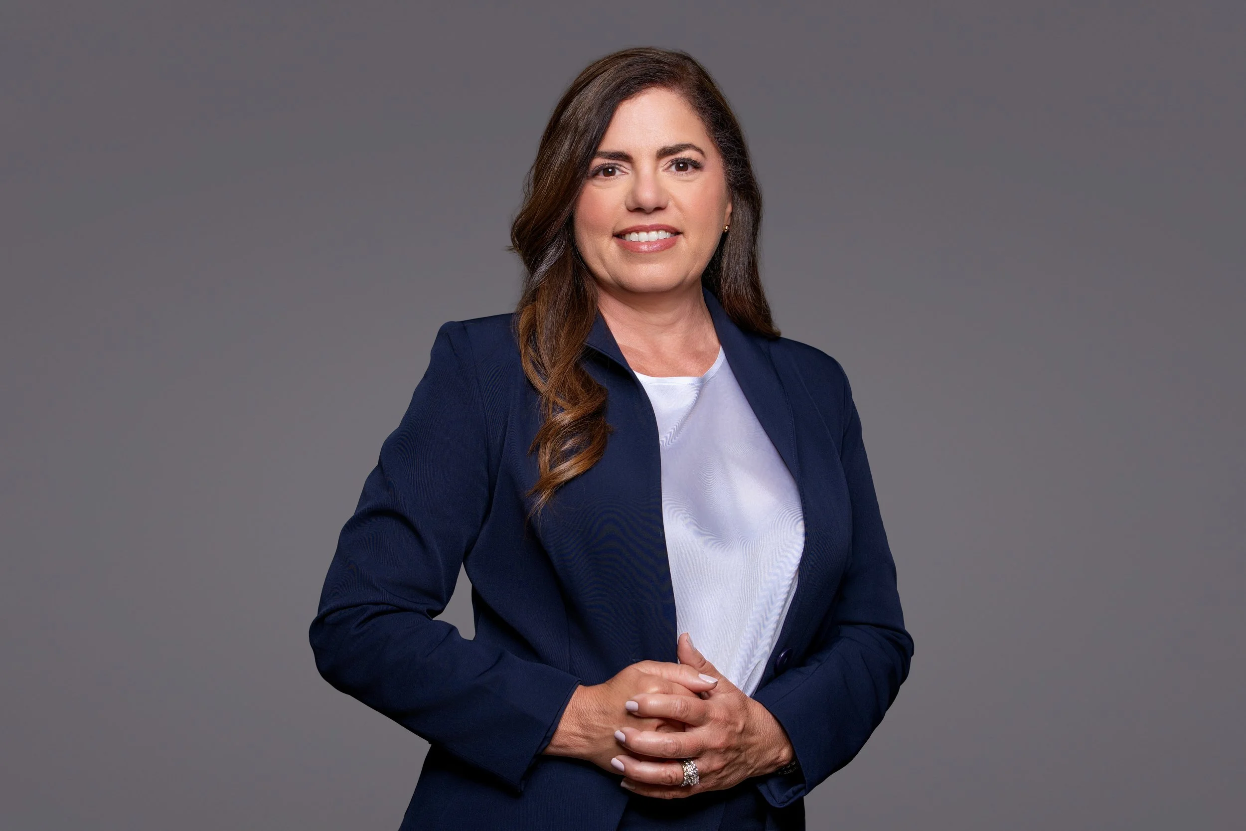 A professional woman with long brown hair, wearing a navy blazer and white top, standing against a plain gray background, smiling with her hands clasped in front of her.
