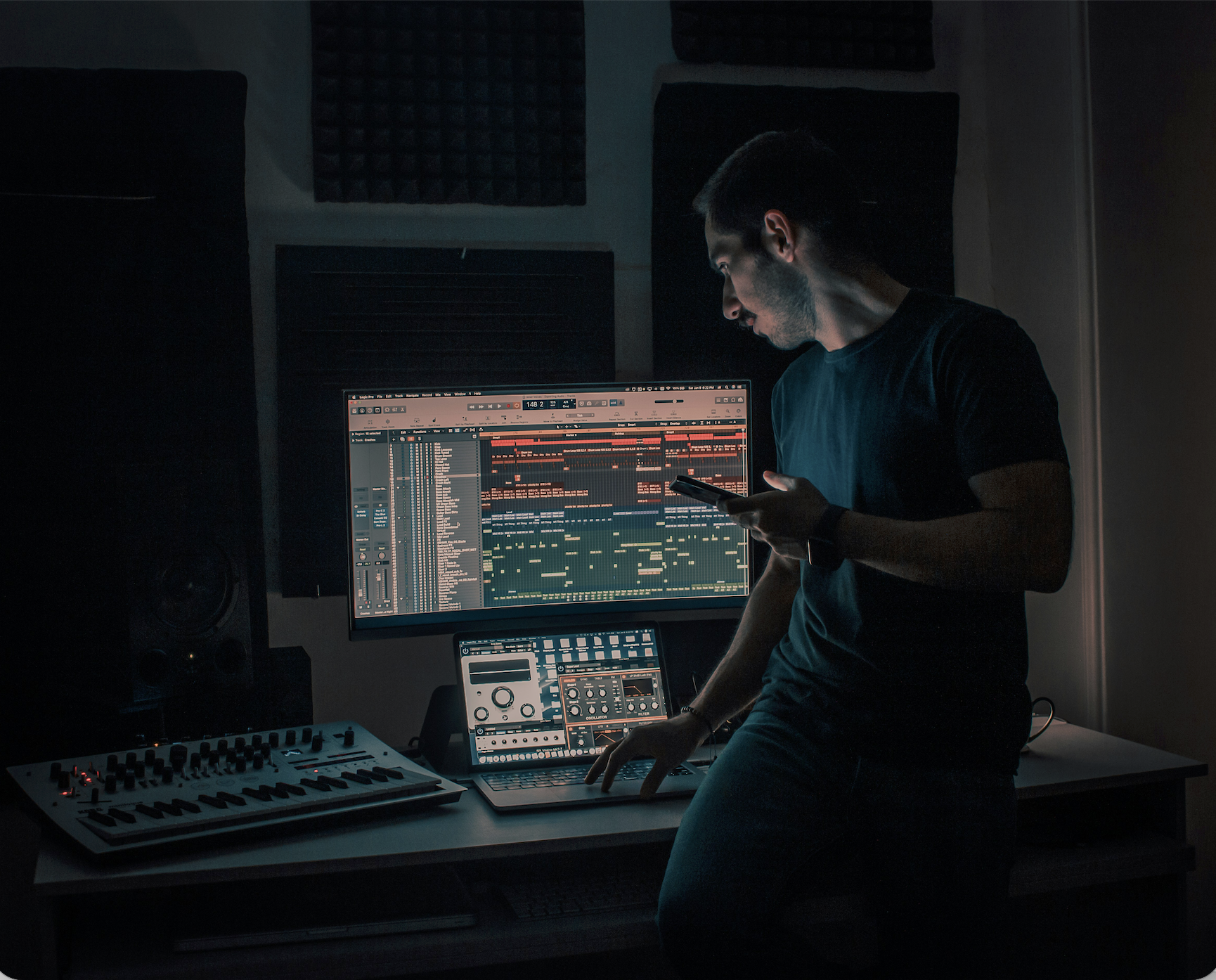 A young man working in a dark music studio, operating a computer with digital audio workstation software on the screen, surrounded by music equipment and soundproofing panels.