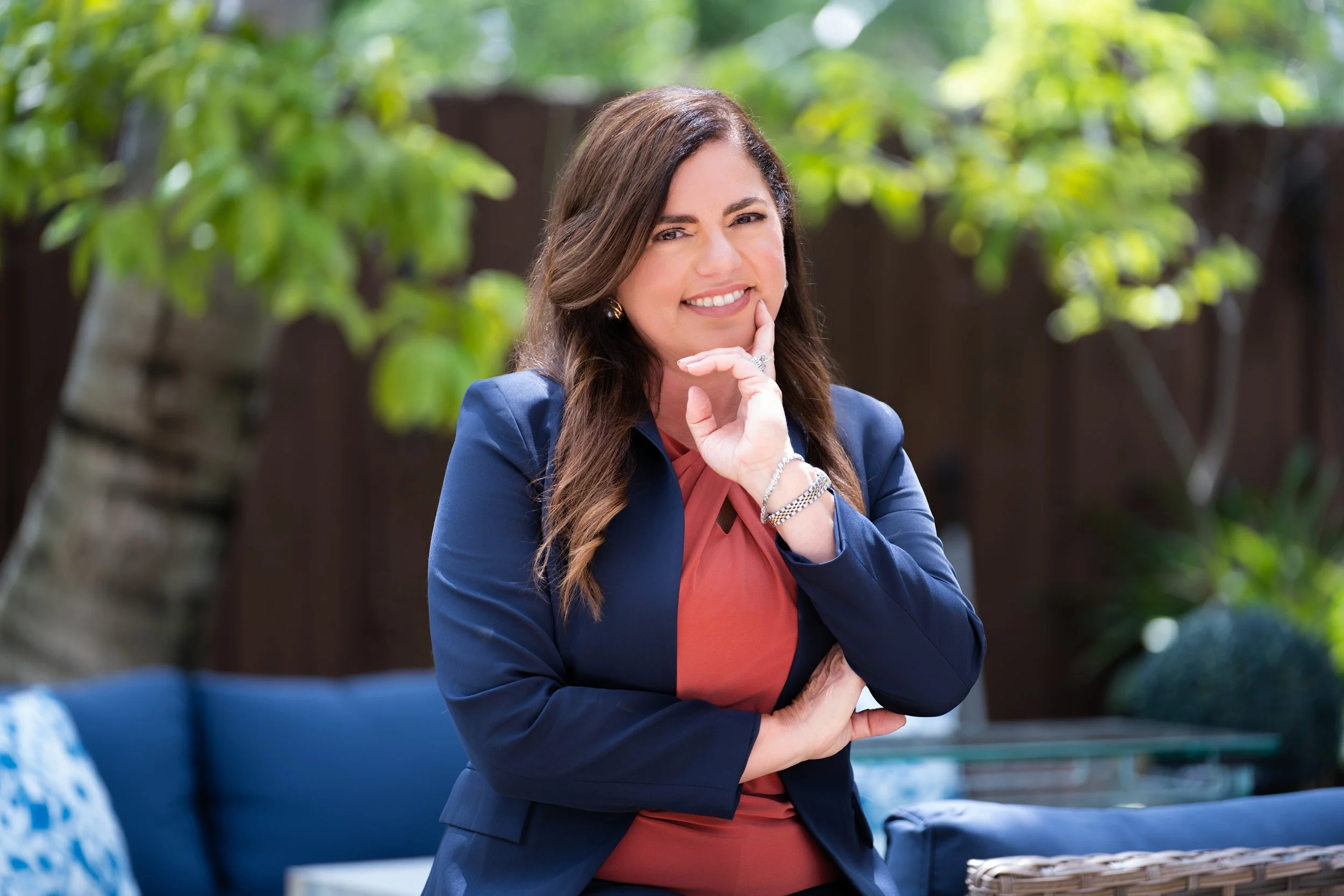 A woman with long brown hair in a blue blazer and orange blouse, posing outdoors with greenery in the background, smiling and resting her chin on her hand.