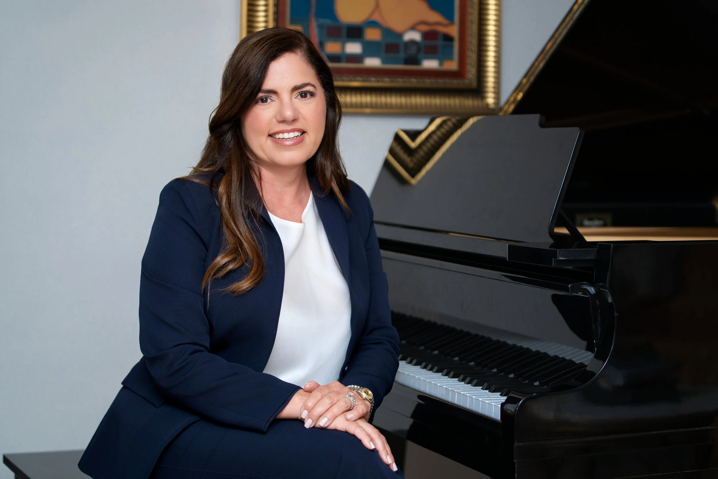 A woman with dark brown hair and light skin, sitting at a black grand piano, wearing a navy blue blazer over a white top, with a framed artwork on the wall behind her.