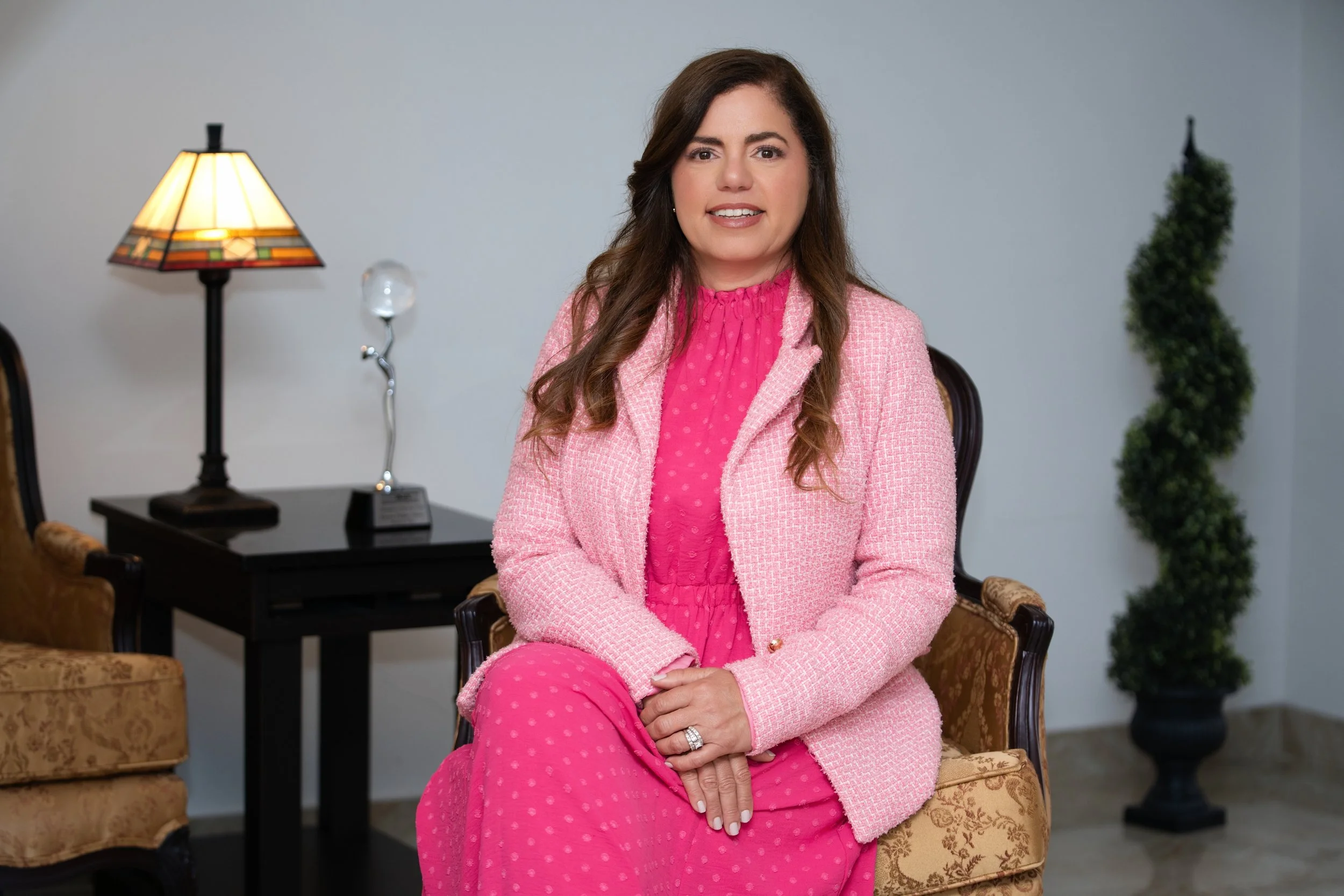 A woman with long brown hair wearing a pink textured blazer and pajamas sits on an ornate chair in a room with classic furniture and a modern background. There is a lamp with a stained glass shade, a trophy, and a tall potted plant visible in the background.