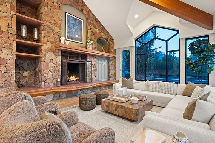 Living room with a large white sectional sofa, a stone fireplace, and a wall of large glass windows showing trees outside.