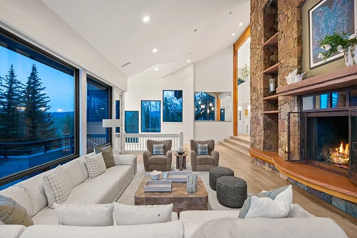 Bright living room with large windows showing a winter landscape, featuring a stone fireplace with a fire burning, white sofa, armchairs, and a cozy home decor.