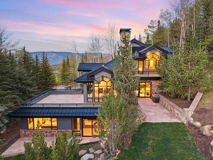 A modern house surrounded by trees with a mountain view in the background, illuminated from inside with large glass windows.