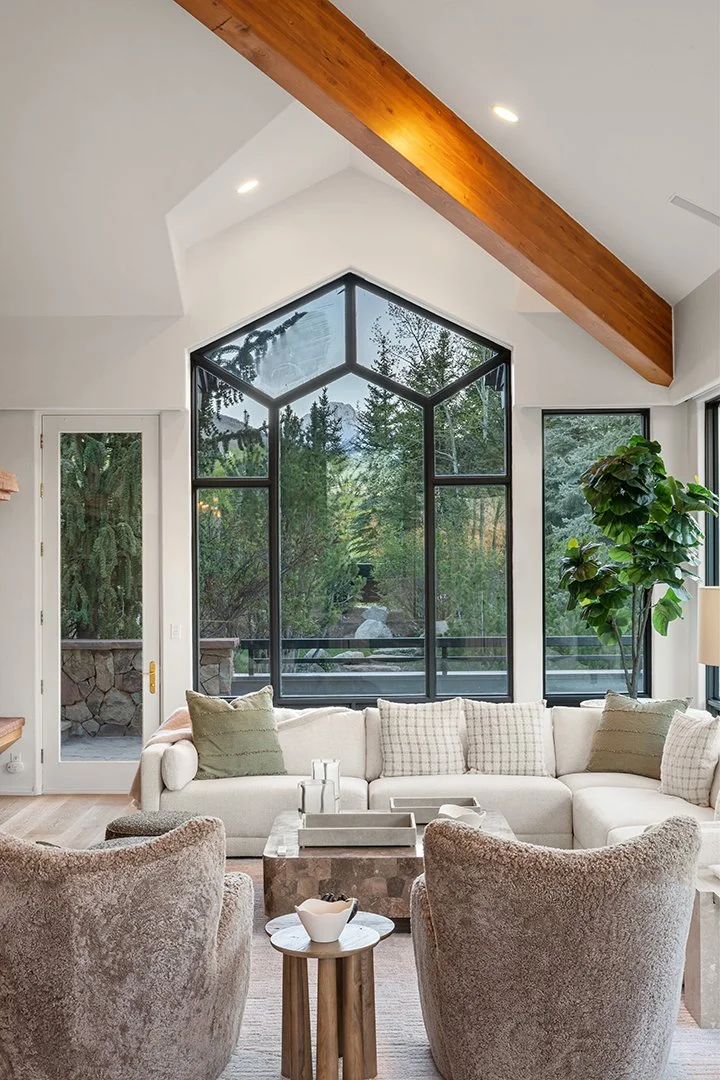 A living room with large geometric windows showing green trees outside, a white sectional sofa with cushions, two beige armchairs, a small wooden side table, and a potted plant in the corner.