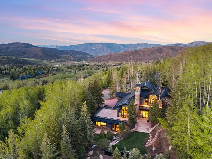 A large modern house surrounded by trees and overlooking a valley with mountains in the background during sunset.