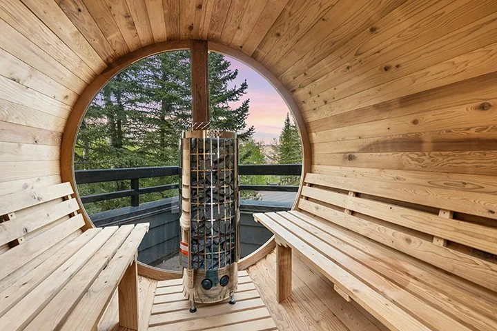Interior of a round wooden sauna room with two wooden benches and a heater in the center, overlooking a forest through a large circular window at sunset.