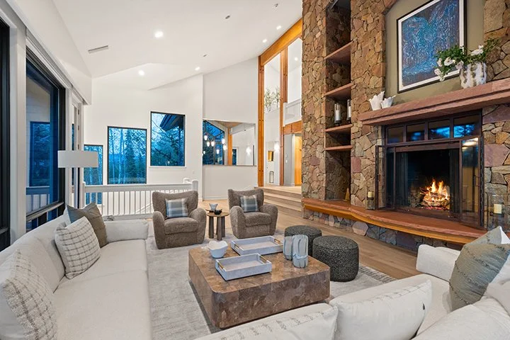 Living room with a white sectional sofa, two armchairs, a stone fireplace, wooden shelving, large windows, and modern decor.