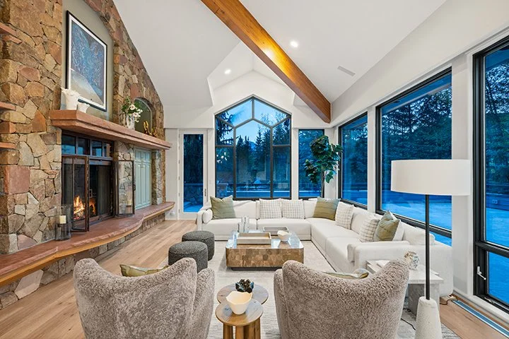 Living room with a stone fireplace, large windows showing trees outside, white sectional sofa, two gray armchairs, wooden coffee table, floor lamp, and some green cushions.