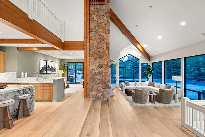 Open-concept living room and kitchen with vaulted ceiling, large windows, and a stone fireplace.