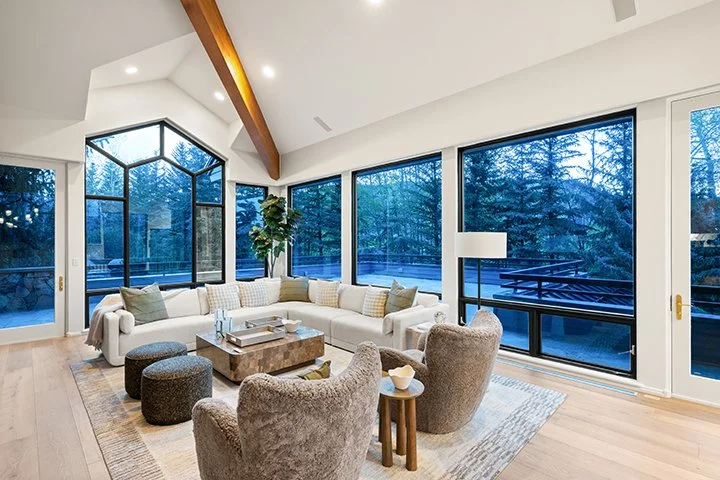 Bright living room with large windows, modern beige sectional sofa, two armchairs, and a wooden coffee table, overlooking a wooded outdoor area.