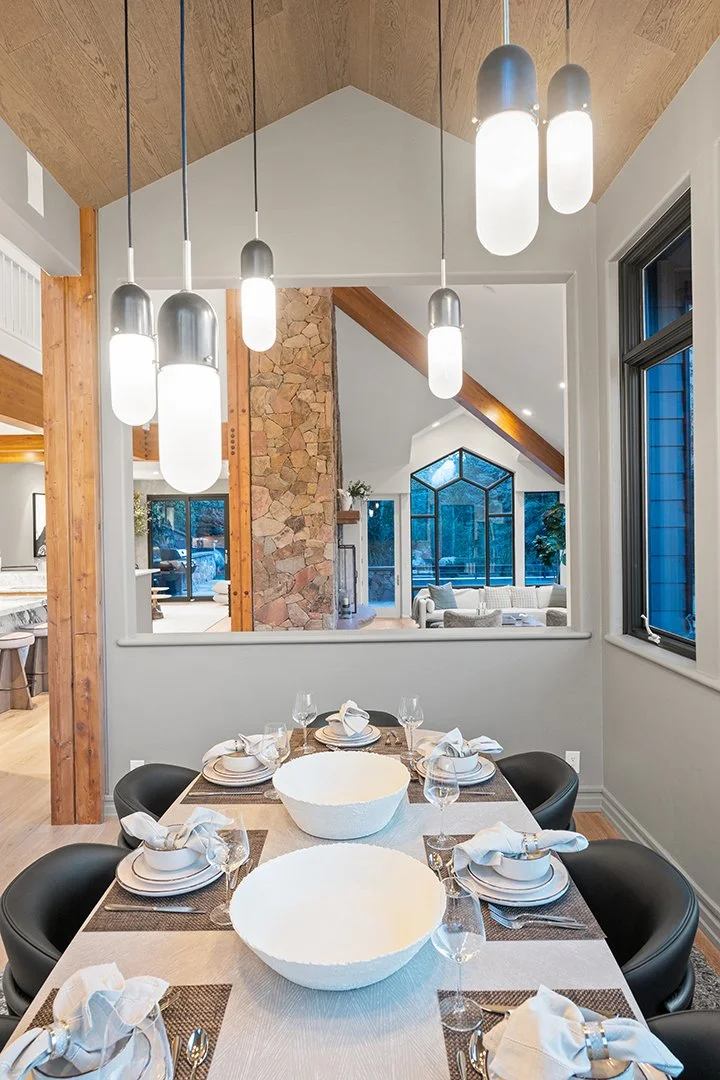 Modern dining room set for six with black chairs, a long table with a light tablecloth, two large white bowls, and table settings including napkins, plates, forks, and glasses. Above are seven pendant lights hanging from a wooden ceiling. A window wa