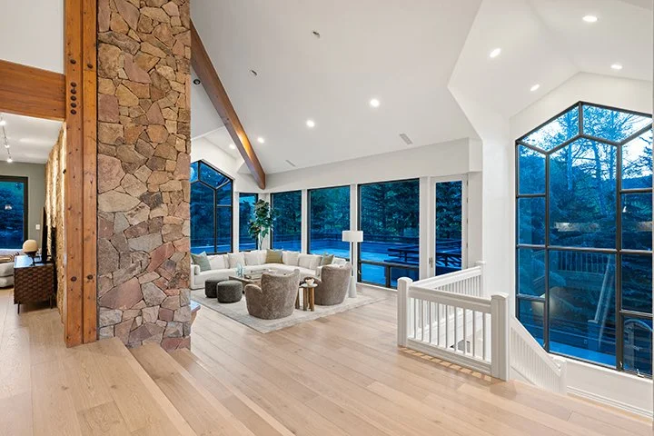 Bright living room with large windows, white walls, wooden beams, stone wall, and modern furniture including a sectional sofa and area rug.