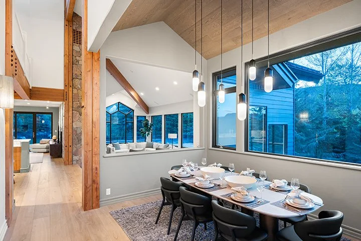 Modern dining room with wooden ceiling, large windows, and a long table set for a meal with plates, glasses, and napkins. Pendant lights hang above the table.