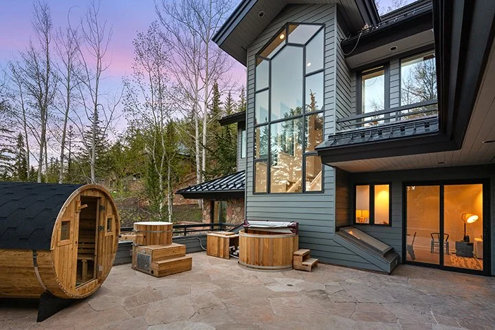 Modern house with large glass windows, wooden hot tubs, and outdoor seating area, surrounded by trees at sunset.