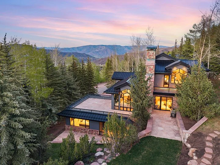 A modern house surrounded by trees with mountains in the background during sunset, with large windows, a stone chimney, and a landscaped yard.