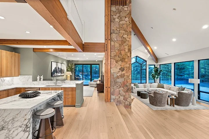 Open-concept living area with kitchen and sitting space, featuring wood beams, a stone pillar, large windows, and modern furniture.