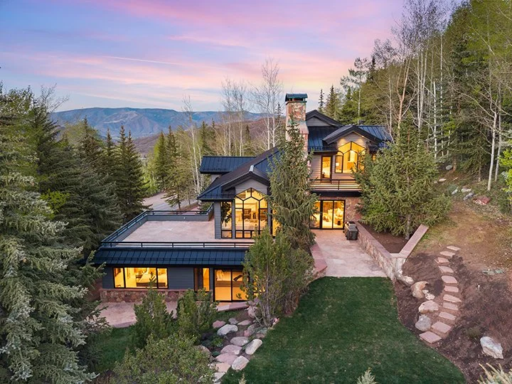 A modern house with large windows and a chimne, situated among trees in a hilly area at sunset.
