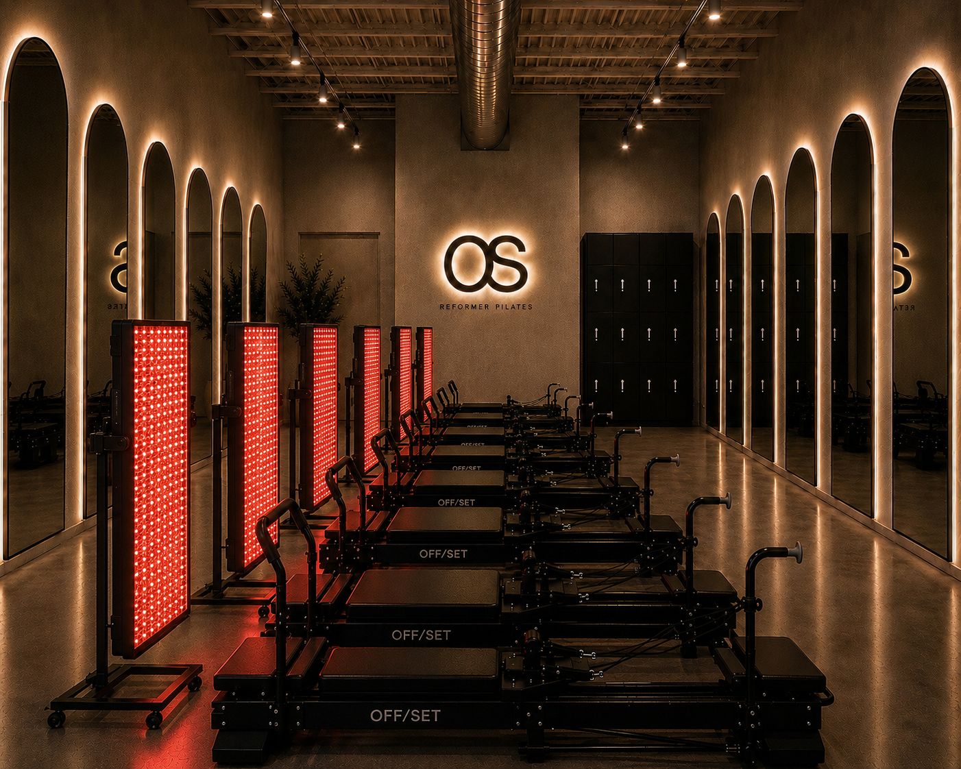 Fitness studio with reformer Pilates equipment, illuminated mirrors along the walls, and a backlit logo that reads 'OS Reformer Pilates' on the wall.