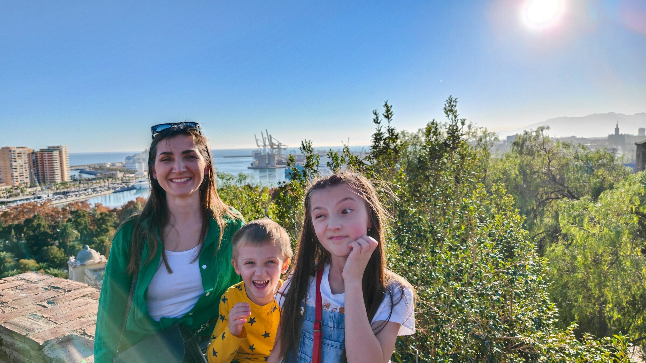 Four people smiling outdoors with cityscape, harbor, and mountains in the background under a bright sun.