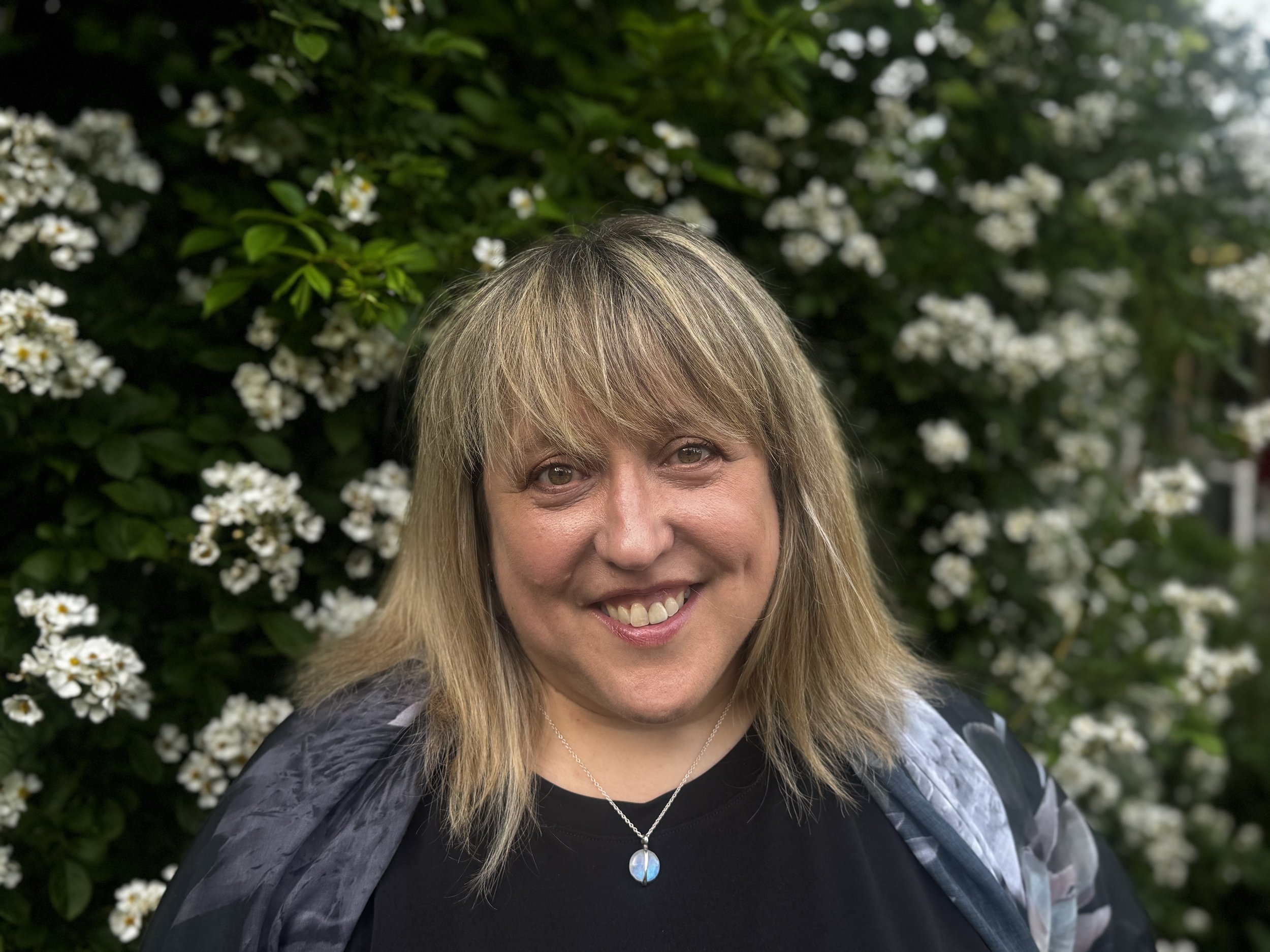A woman with light brown hair and bangs, smiling outdoors in front of a green bush with small white flowers, wearing a black top, a patterned jacket, and a necklace with a blue and silver pendant.