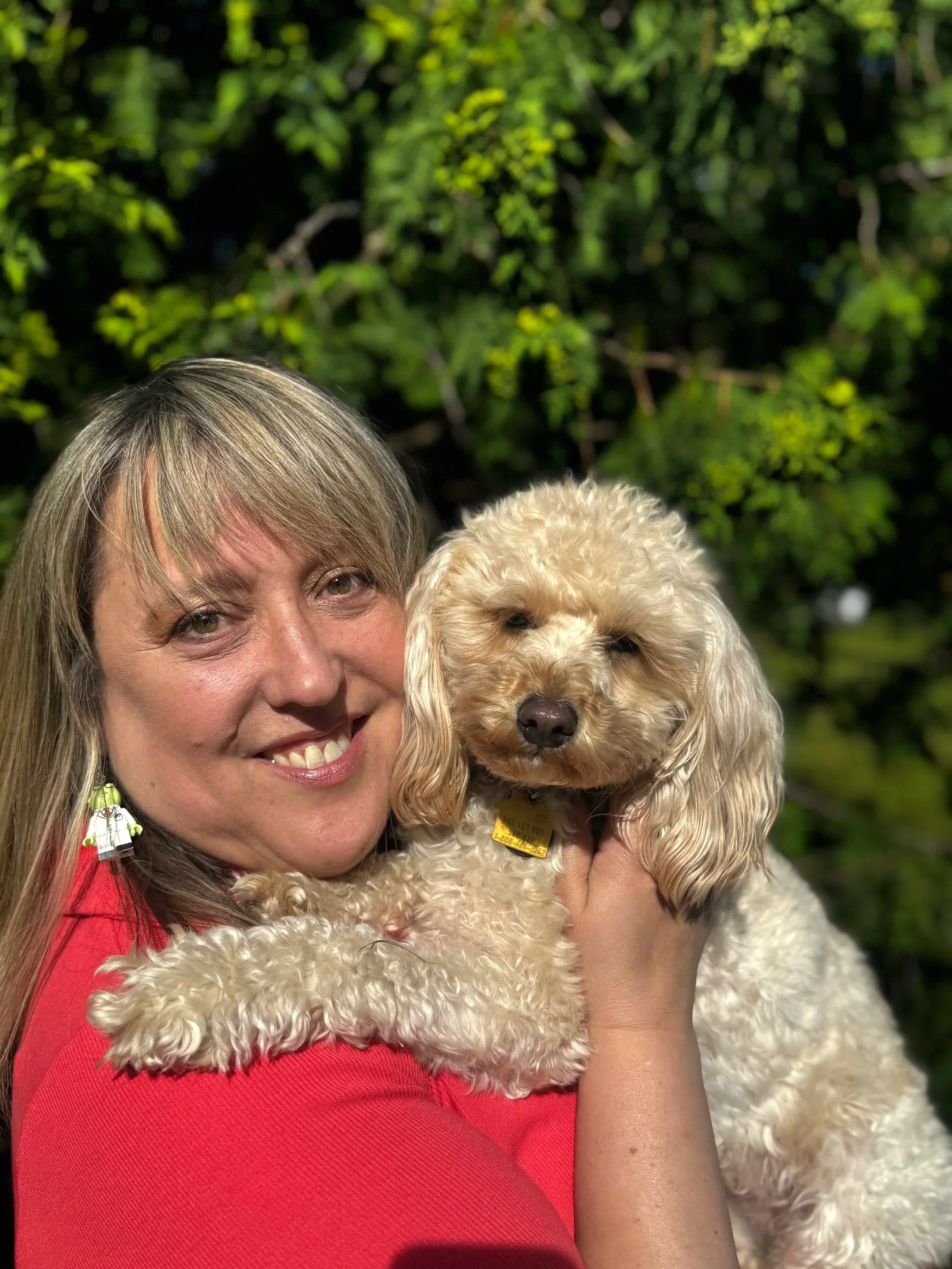 Woman with blonde hair smiling, holding a poodle dog outdoors at night with green foliage in the background.
