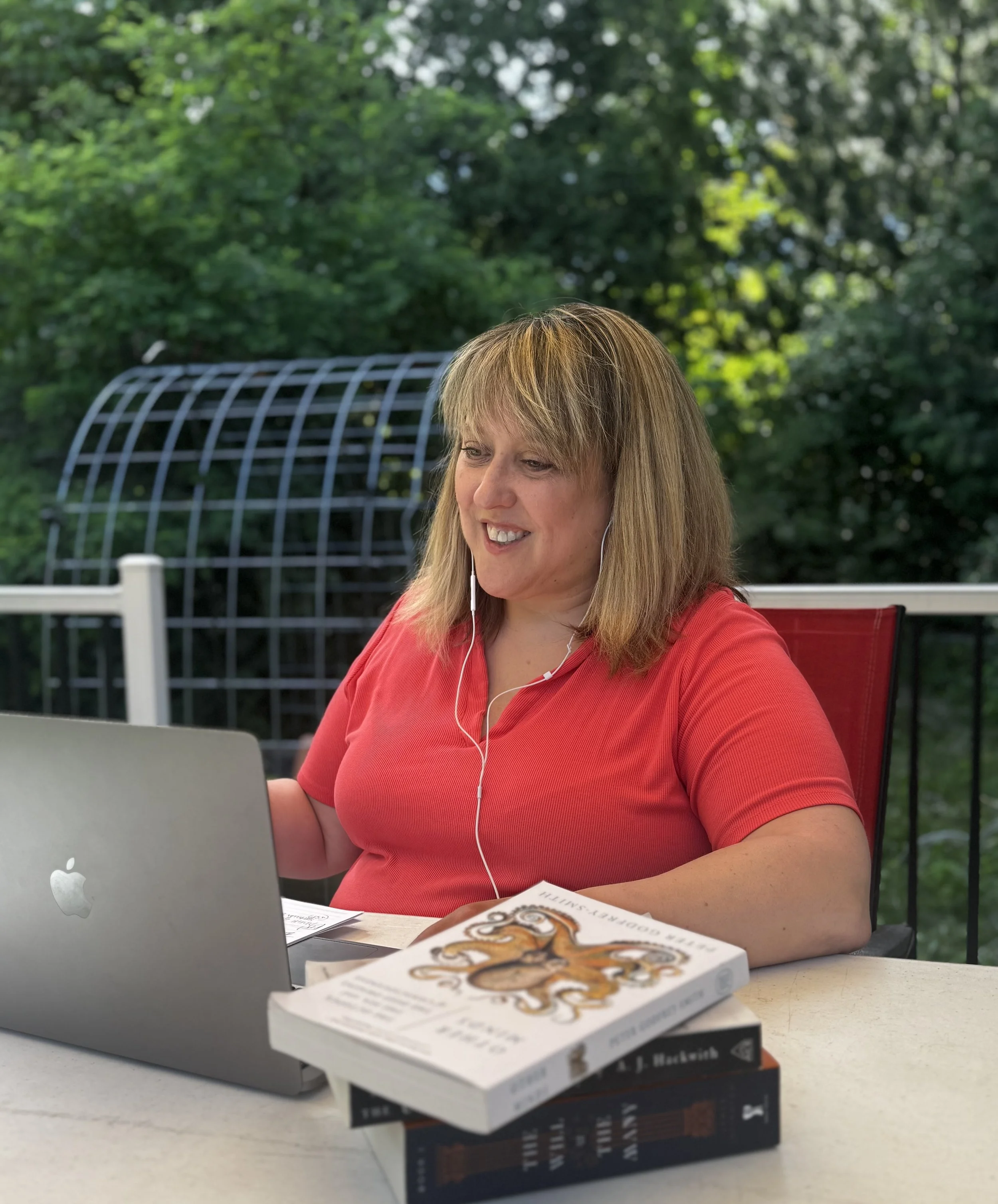 A woman with blonde hair wearing a red shirt, sitting at a white outdoor table, working on a laptop and listening to headphones. There are books, including one titled 'The Book of Joy,' on the table in front of her. The background features green foliage and an outdoor covered structure.