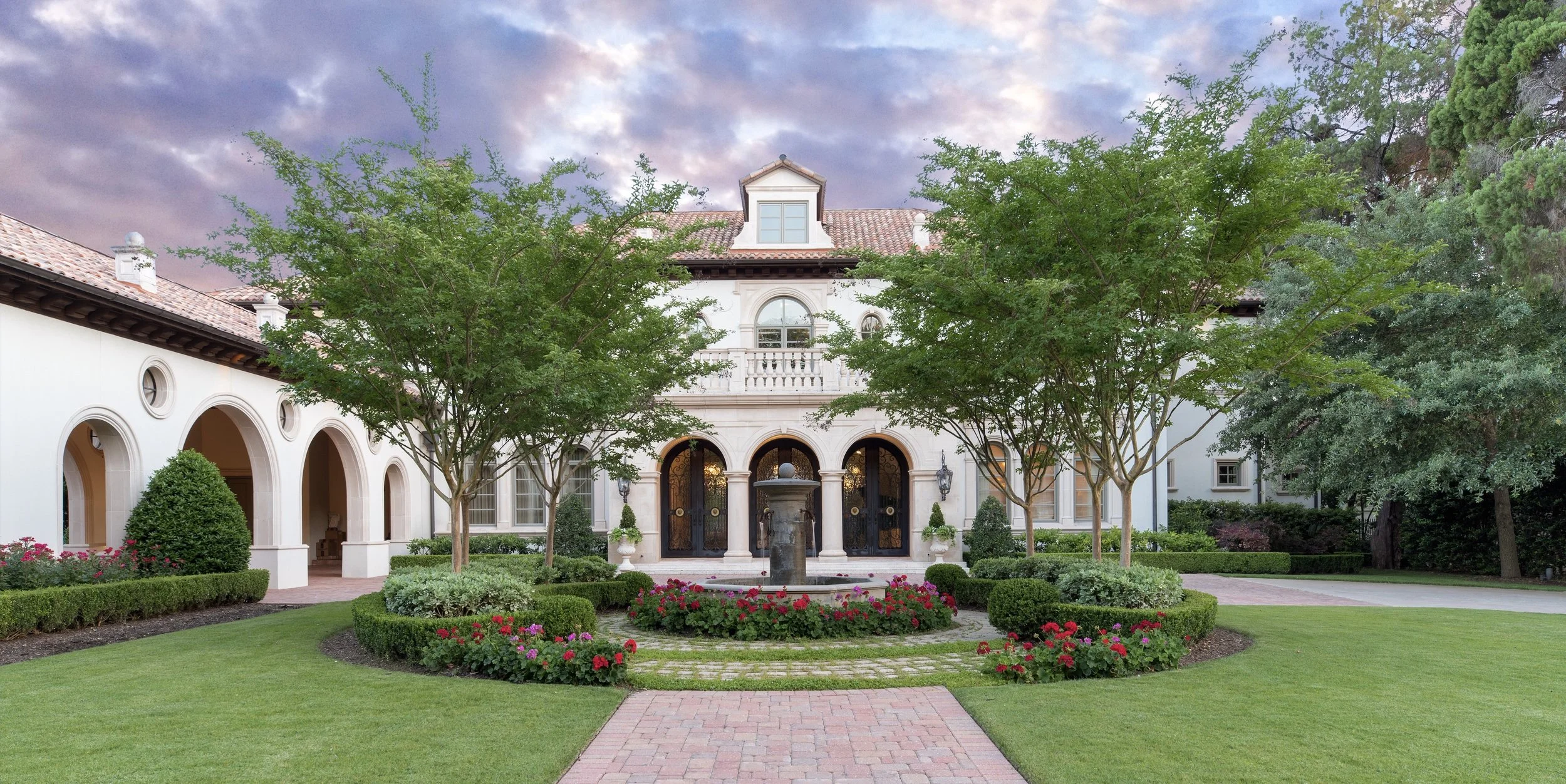A luxurious white house with an arched entrance, surrounded by a landscaped garden with green trees, colorful flowers, and a fountain, under a partly cloudy sky during the daytime.