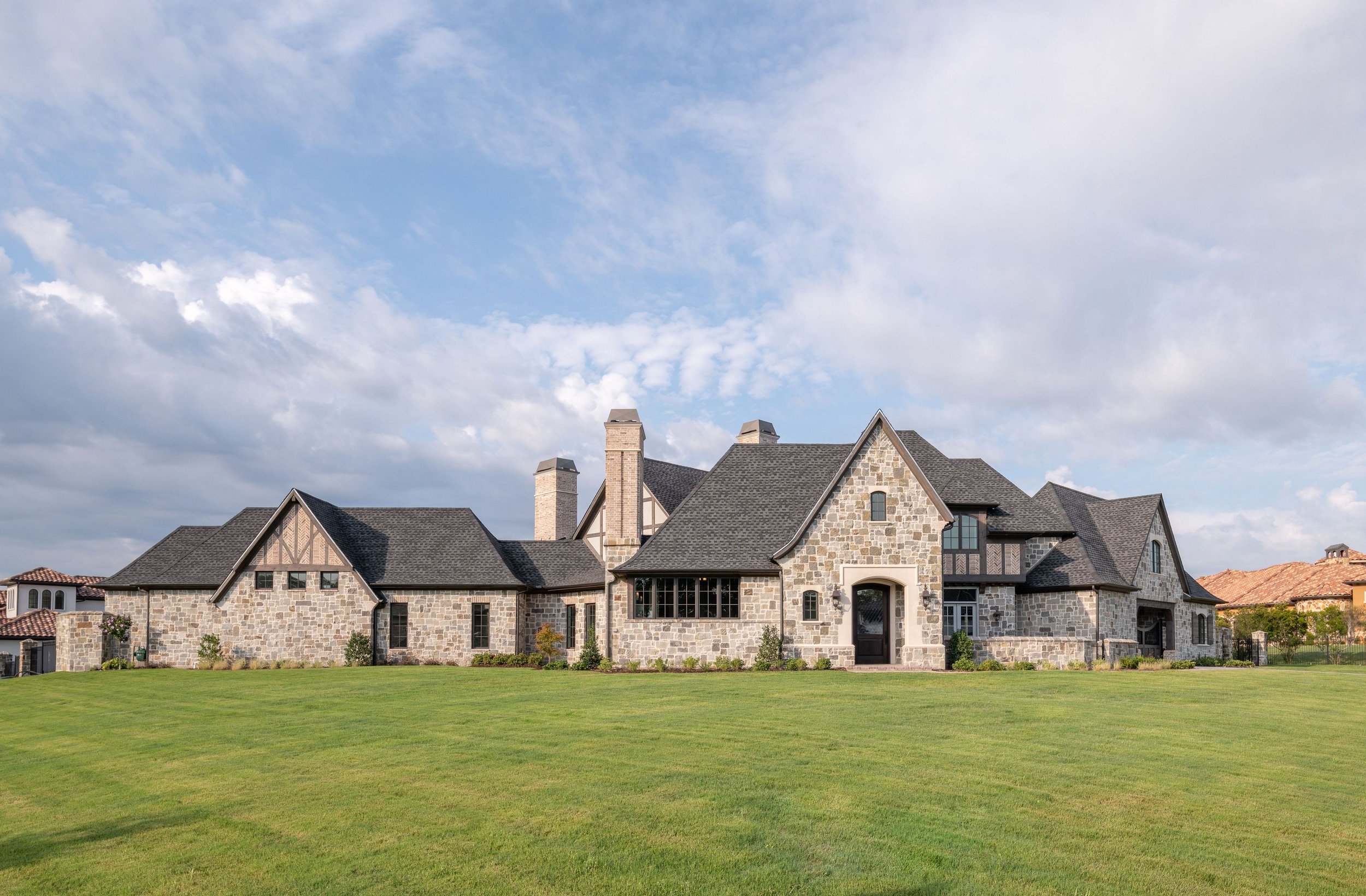 Large stone and brick house with multiple chimneys, dark roof, front yard with green grass, under partly cloudy sky.