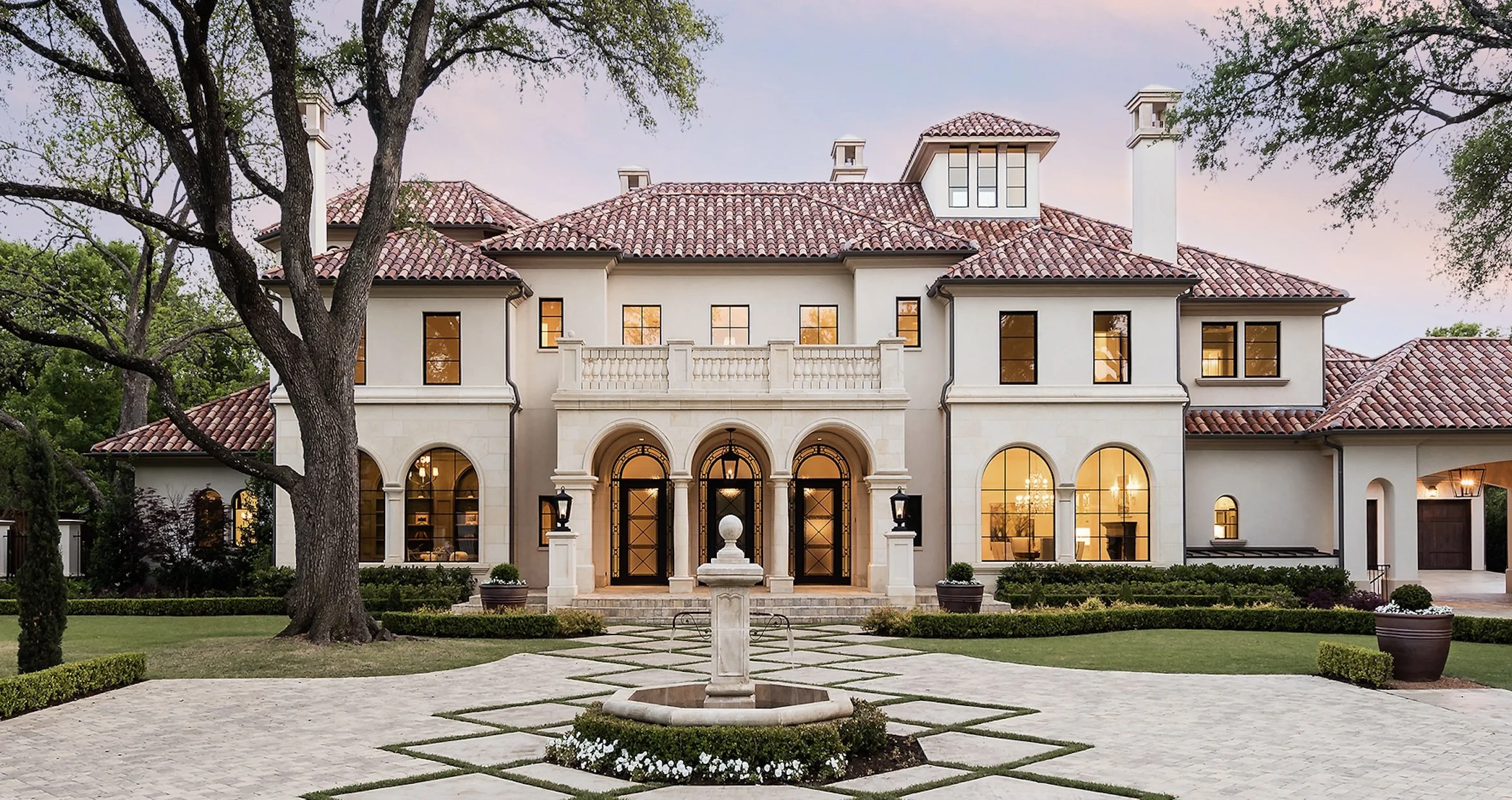Large elegant mansion with a tiled roof, arched windows, and a grand entrance, surrounded by a manicured garden with pathways, potted plants, and mature trees, under a pink and blue evening sky.