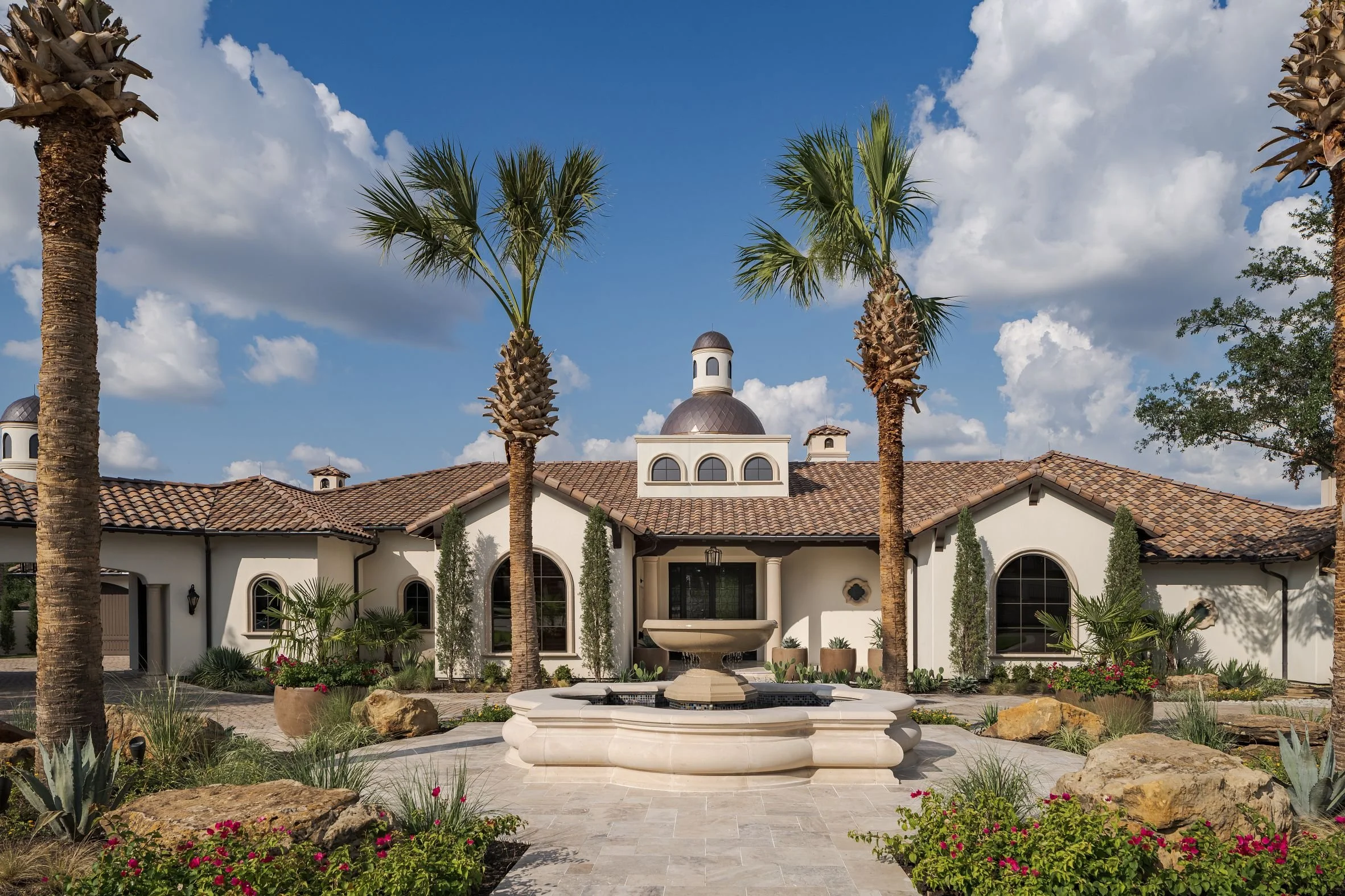 A luxurious house with a tiled roof, surrounded by palm trees and lush landscaping, with a fountain and cloudy sky in the background.