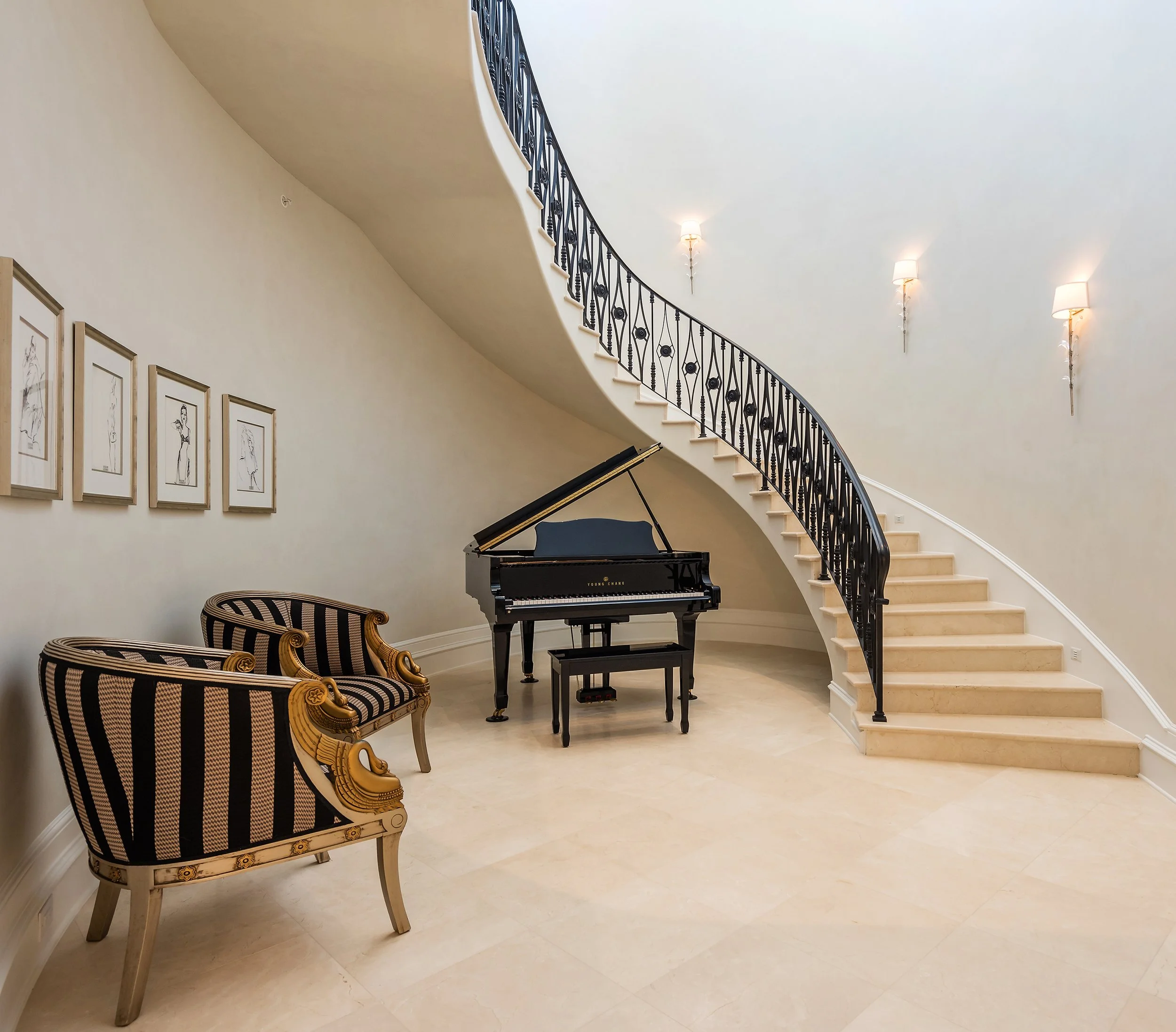 Elegant interior featuring a grand staircase with a black wrought iron railing, a black grand piano, classical style armchairs with gold accents, and framed line drawings on the wall, illuminated by wall sconces.
